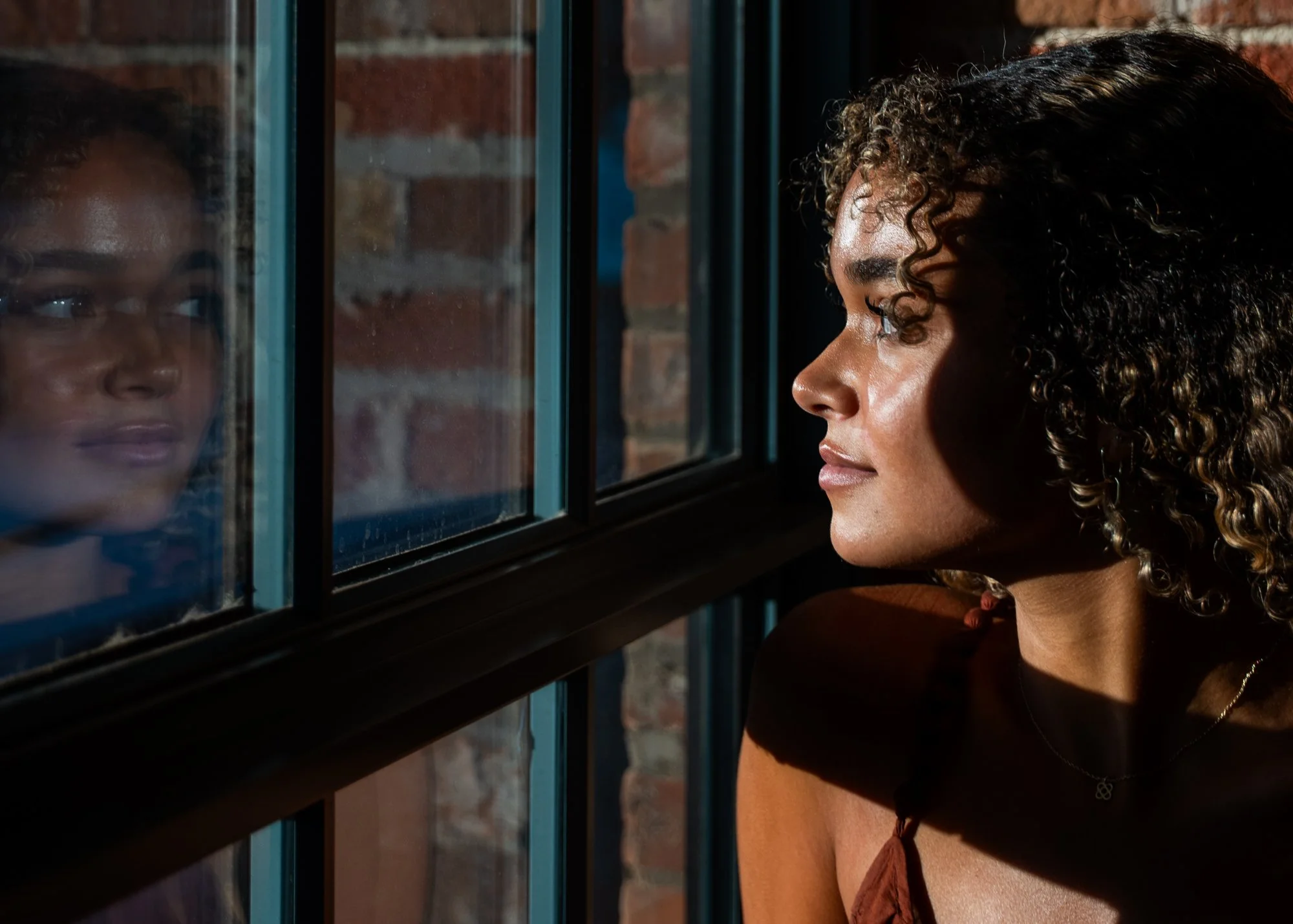 A woman with curly hair looking out the window, her reflection visible on the glass, by a brick wall.