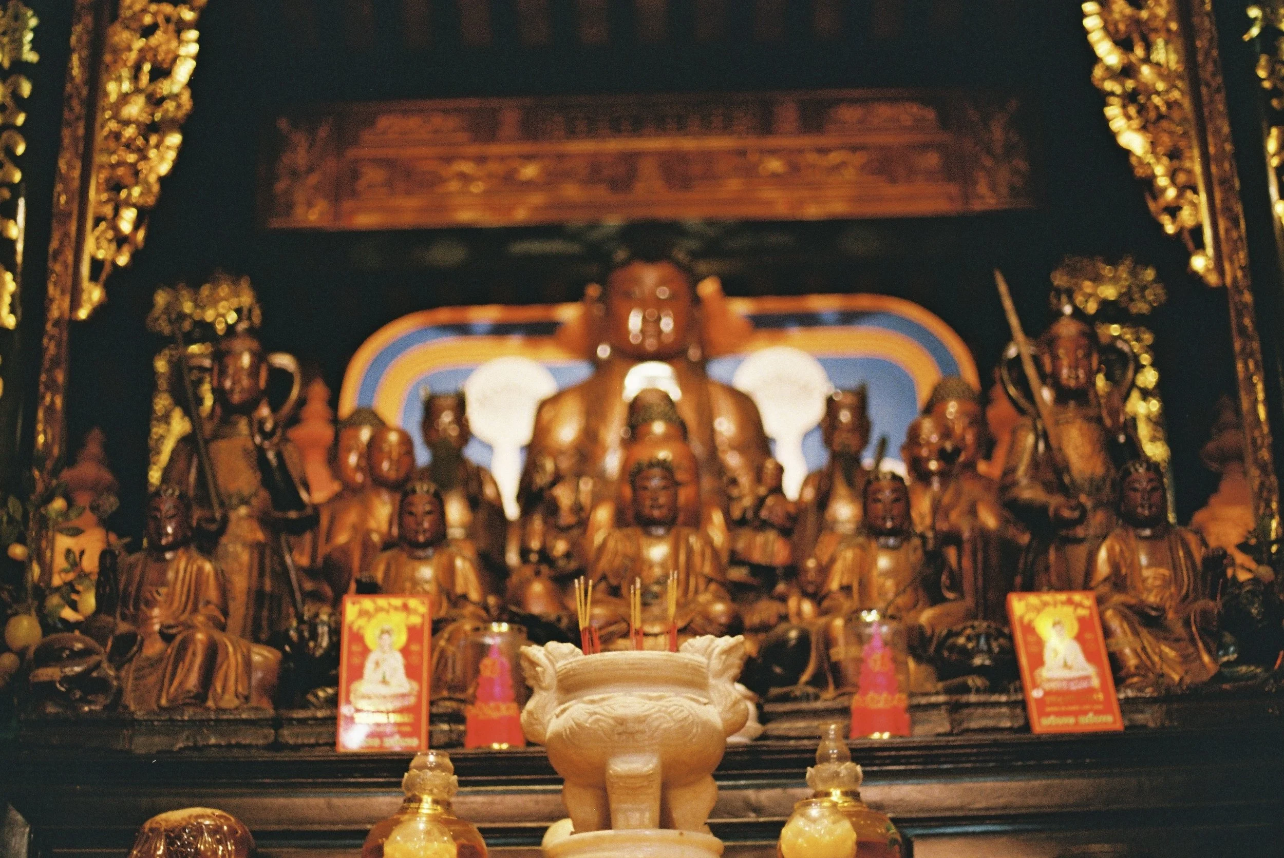 Indoor Buddhist shrine with multiple gold and dark-colored statues of Buddhas and deities, decorated with gold accents, flowers, and offerings, with ornate wooden carvings and vibrant background artwork.