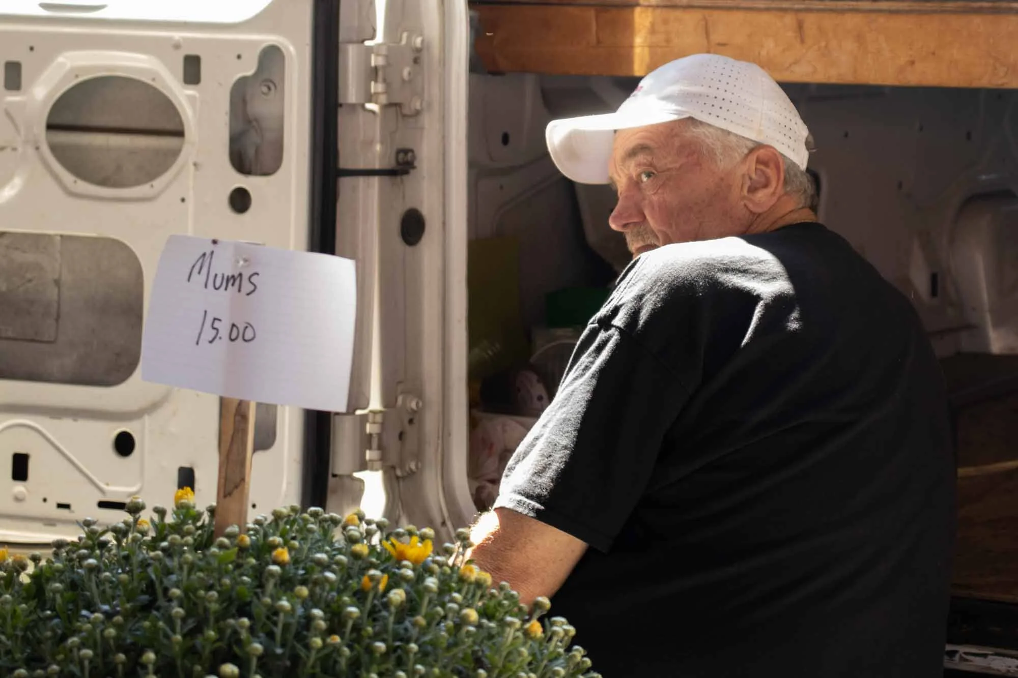 A man wearing a white baseball cap is sitting next to a washing machine with a handwritten sign reading 'Mums 15.00'. The man appears to be selling flowers visible in the foreground.