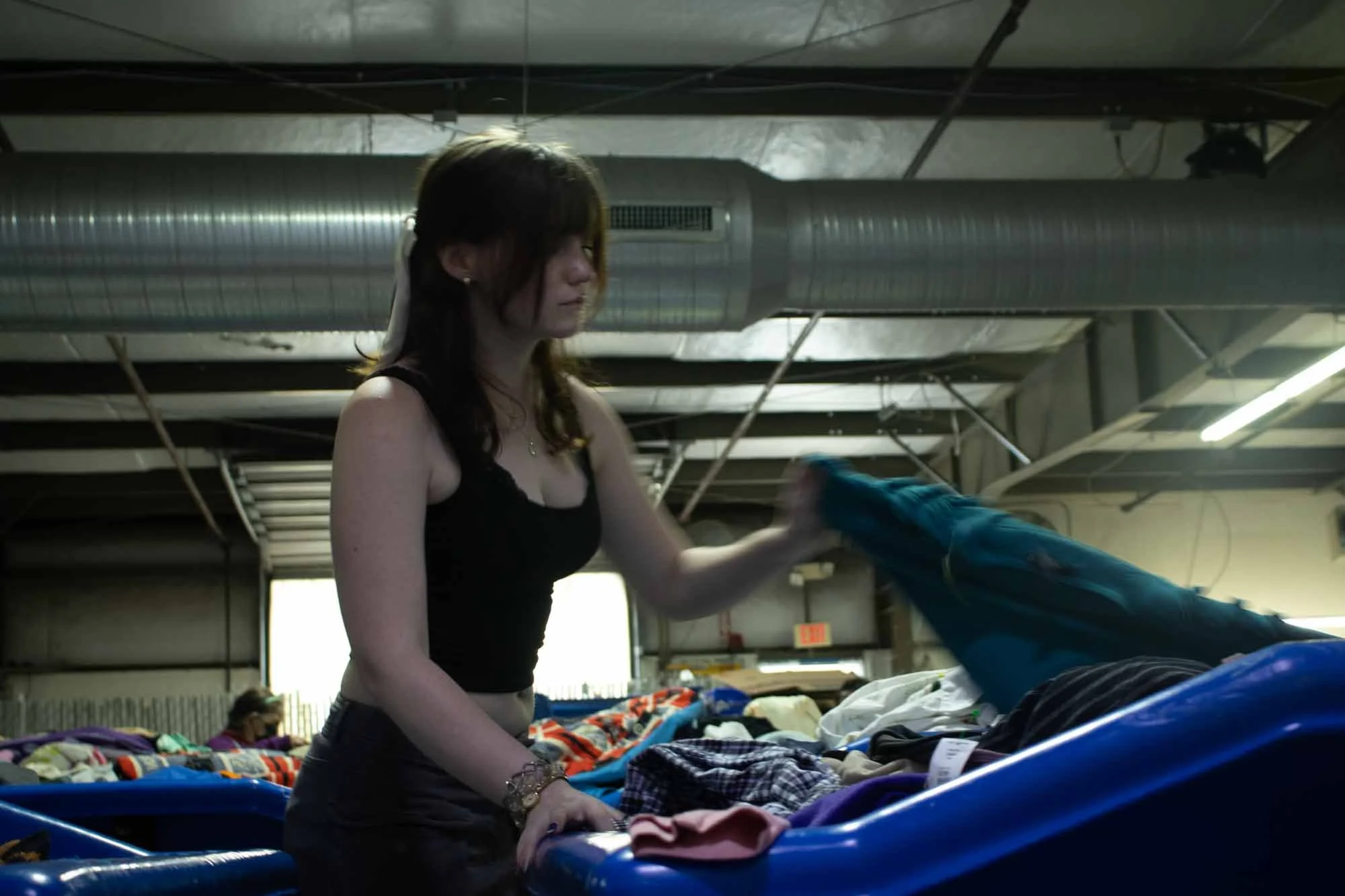 A woman in a black sleeveless top and jewelry is sorting clothes on a table at a thrift store or clothing donation center, with racks of folded and hanging clothes in the background.