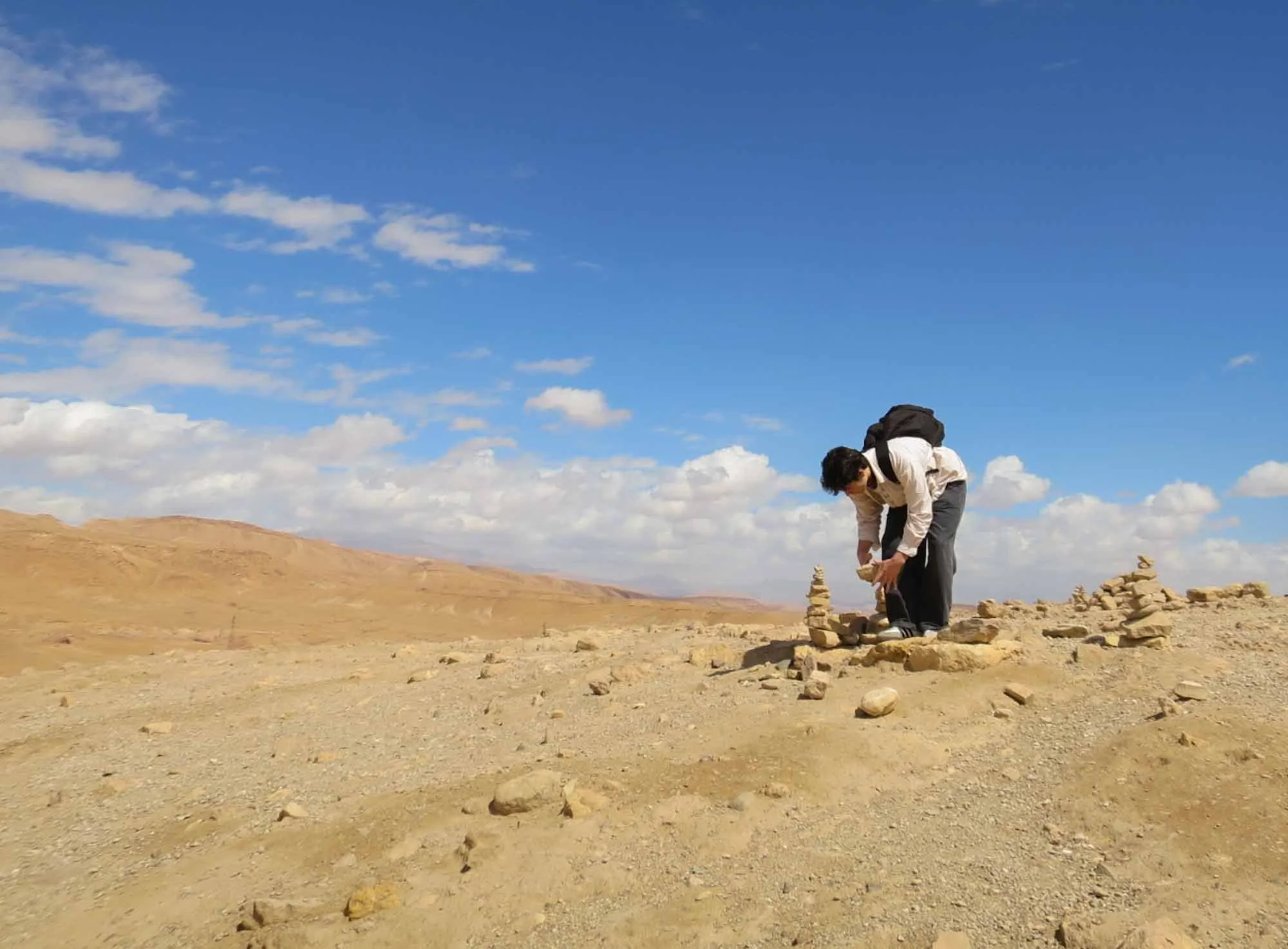 A person with dark hair, wearing a white shirt and gray pants, bending over to stack rocks into small cairns on a dry, grassy plain with hills in the background under a partly cloudy blue sky.