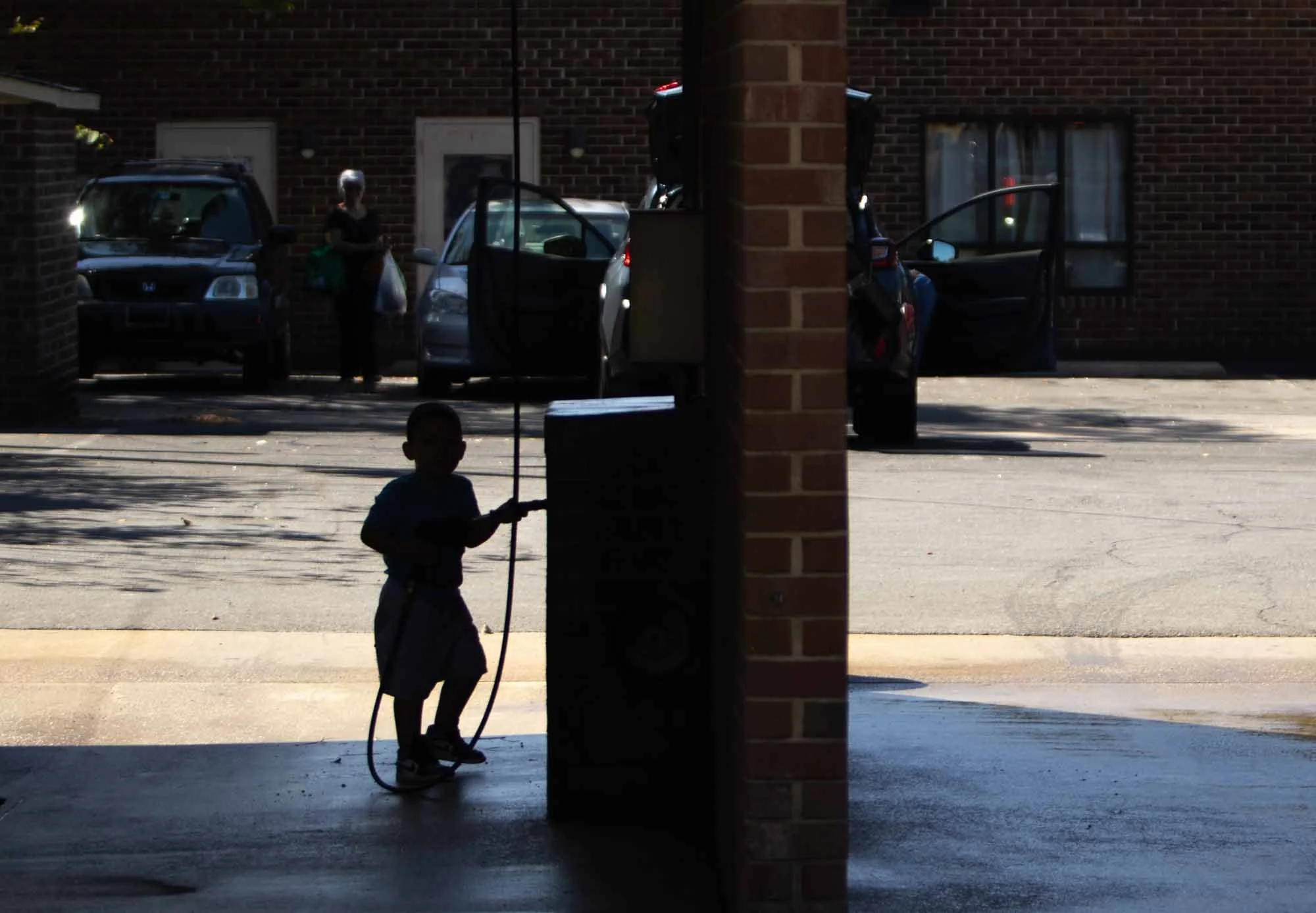 Silhouette of a young boy holding a garden hose standing on the porch, with a view of parked cars and a brick house in the background.
