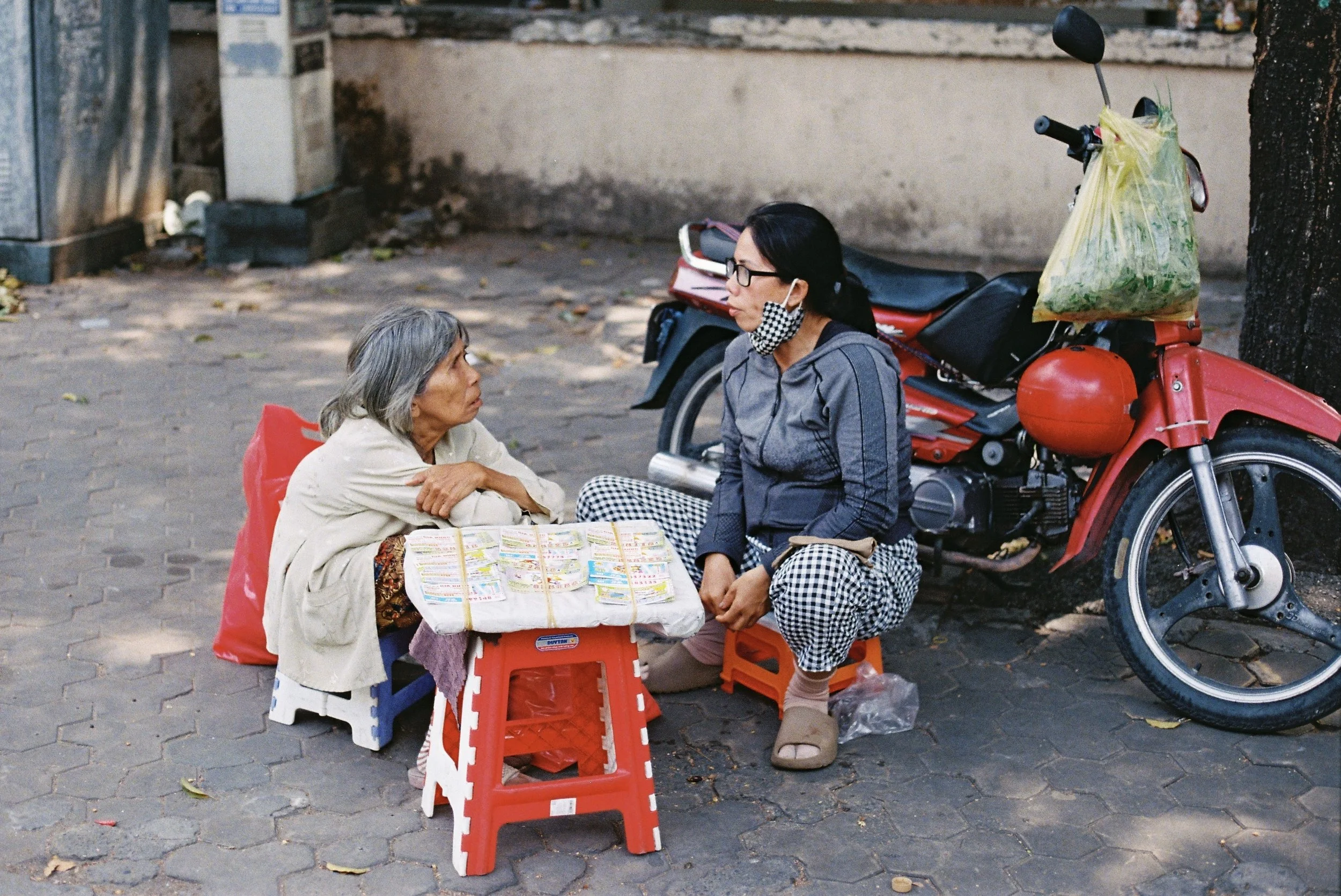 A woman wearing glasses and a black-and-white checkered mask is sitting on an orange plastic stool, looking at an elderly woman with gray hair, who is seated on the ground with a red plastic bag beside her. They are outdoors on a cobblestone street, 