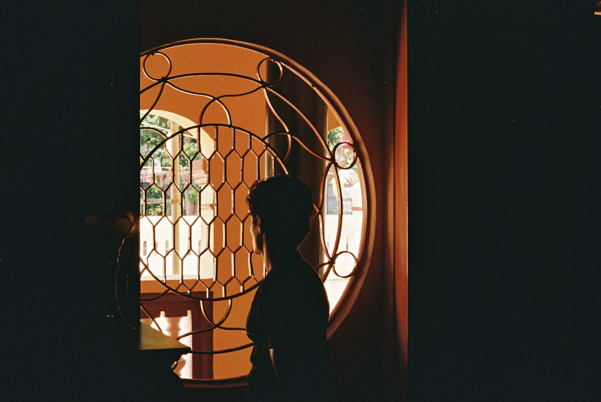Silhouette of a person with short hair looking out a decorative iron window with geometric patterns, sunlight outside illuminating the scene.