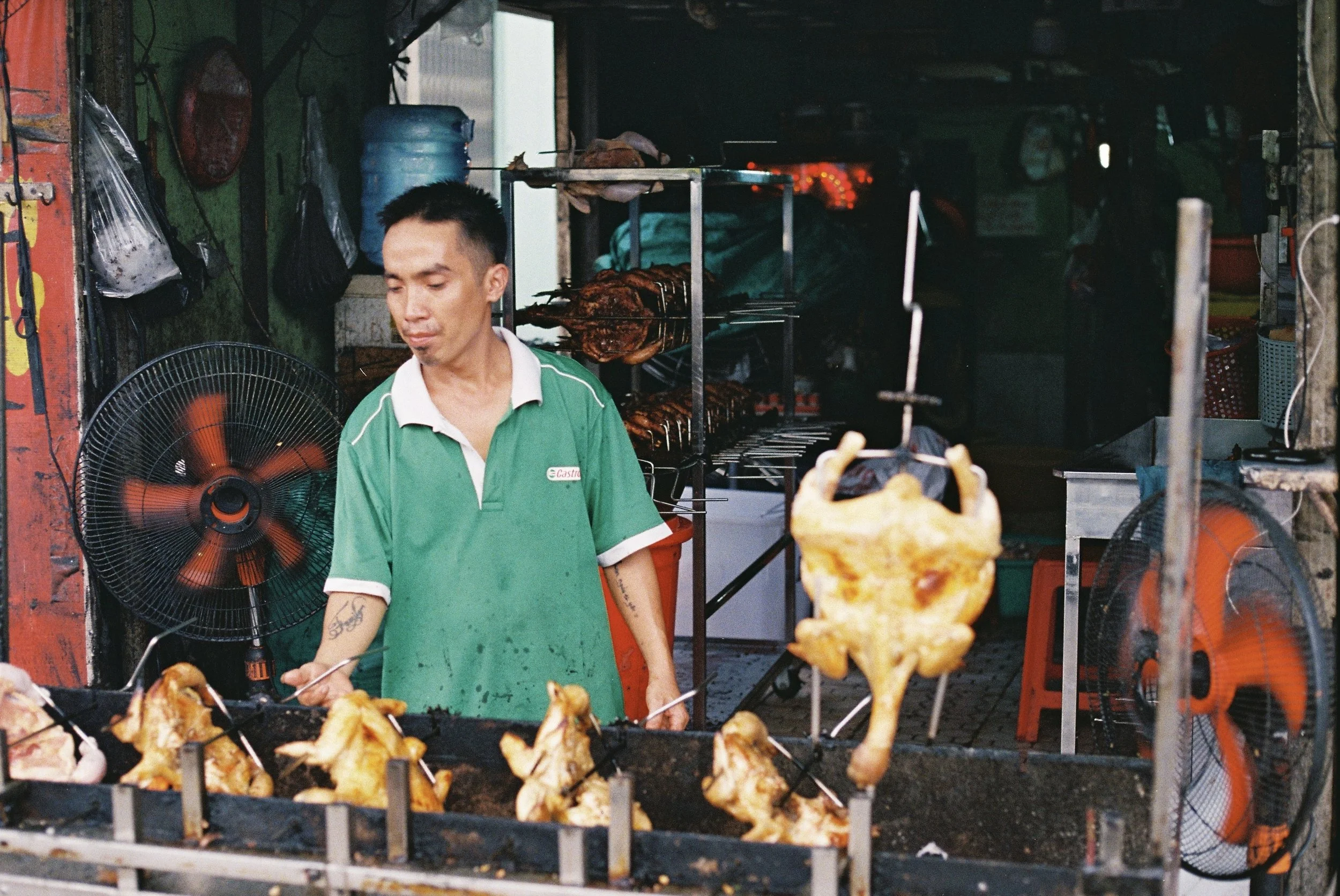 A man wearing a green shirt is grilling chicken pieces on skewers at a street food stall. There are two electric fans and a water jug in the background, along with various tools and supplies in a small, open-front setup.