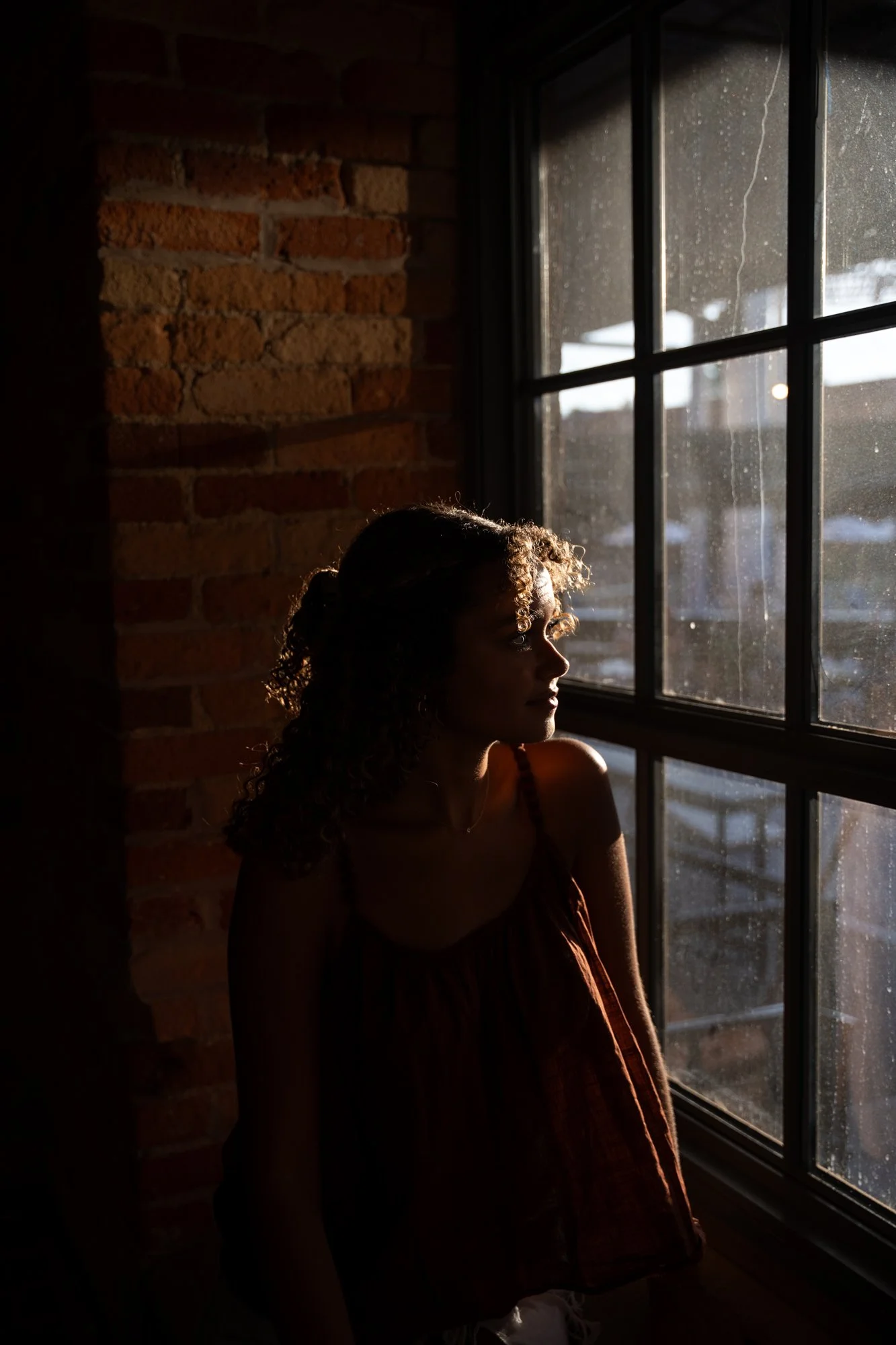 A woman with curly hair looking out a rain-streaked window in a dimly lit indoor space with exposed brick wall.