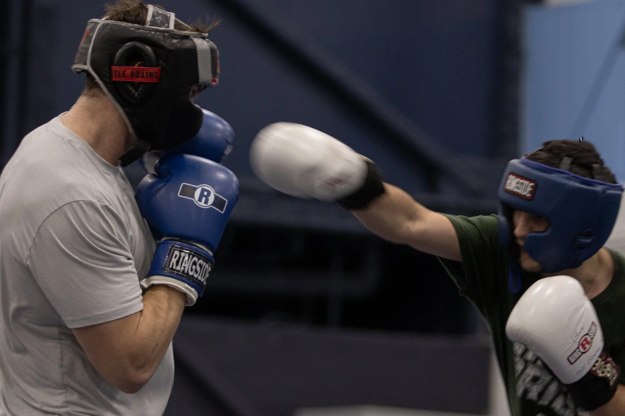 Two people in boxing gear sparring in a gym, wearing protective headgear and gloves, delivering punches to each other.