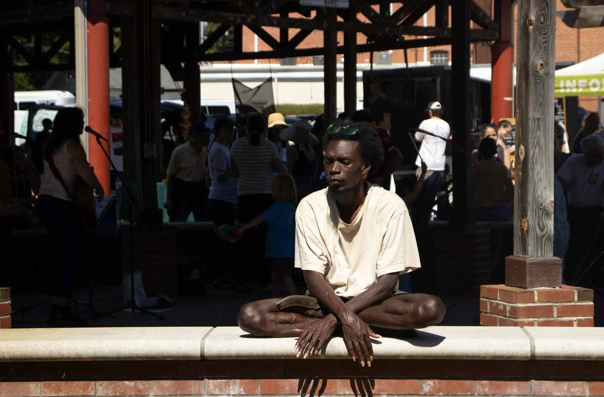 A man with dark skin, black curly hair, and sunglasses resting on his head, seated cross-legged in a meditative pose outdoors in front of a wooden structure, with a crowd of people in the background.