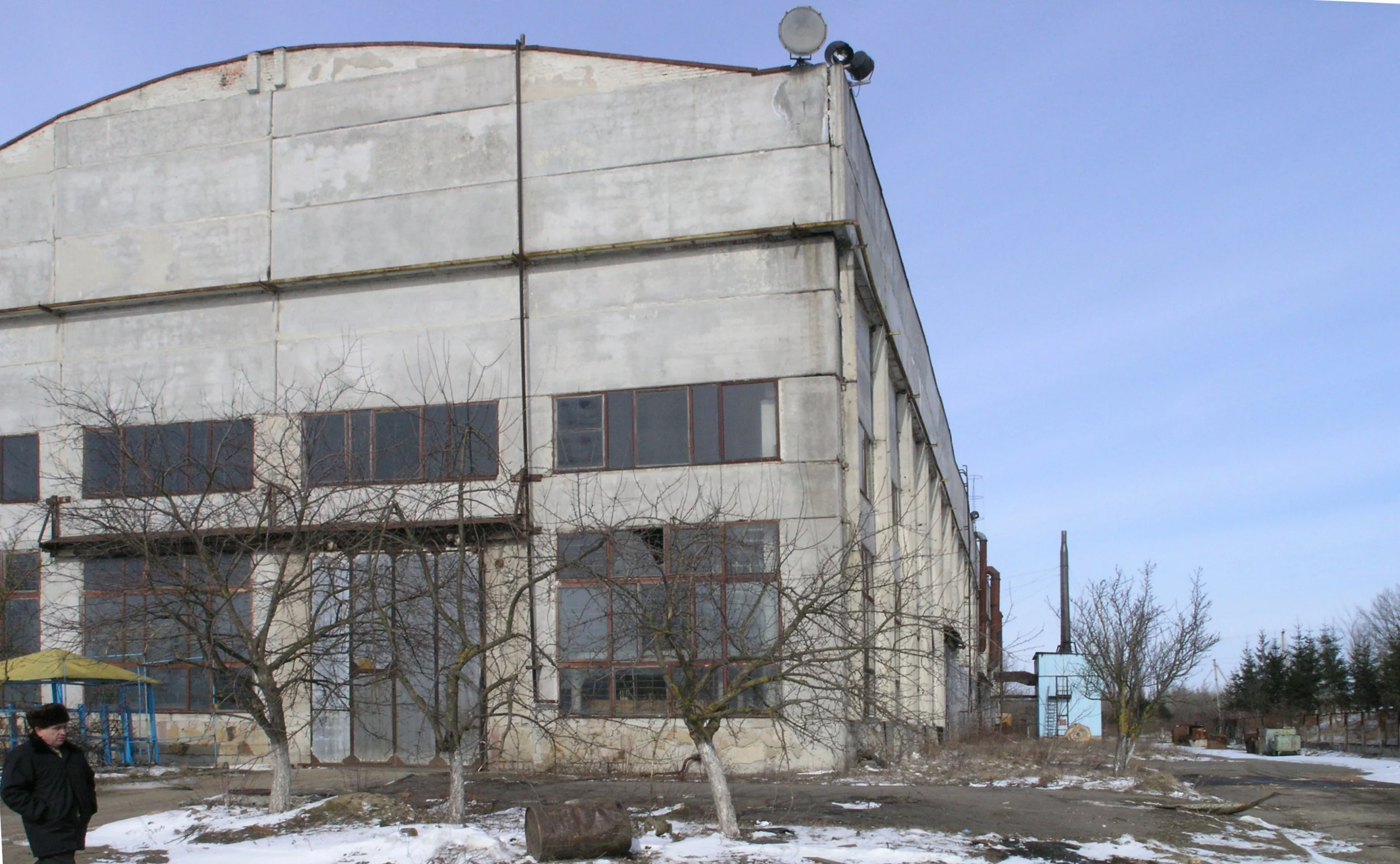 An old, weathered multi-story building with a faded, peeling facade and rusted window frames. In front, there are leafless trees, patches of snow on the ground, and a person wearing a dark jacket walking past. The sky is clear and blue.