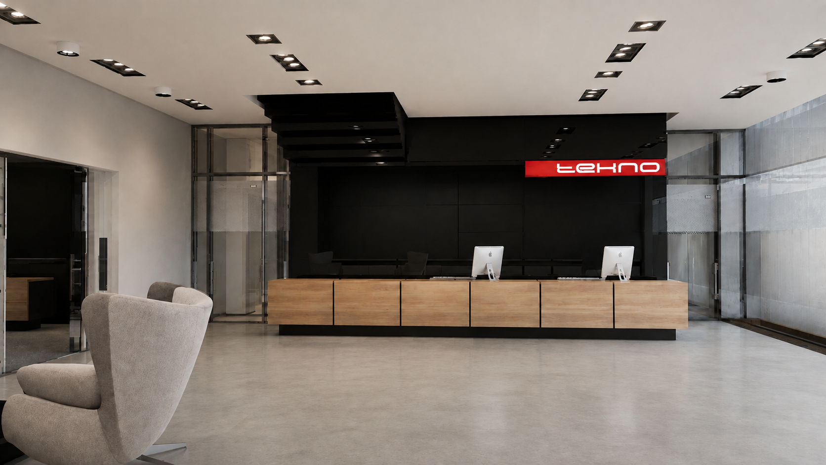 Modern tech company's reception desk with two Apple computers, a beige armchair in the foreground, and a black wall with a partial red sign that reads 'tehno' behind the desk.