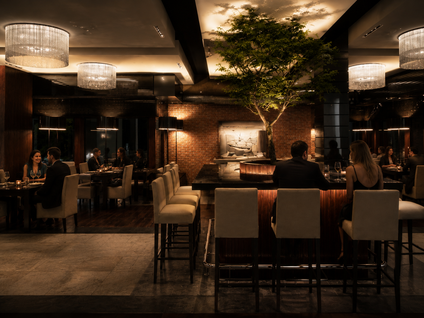 Interior of an upscale restaurant with a central tree, brick wall, elegant chandeliers, and patrons dining and chatting.