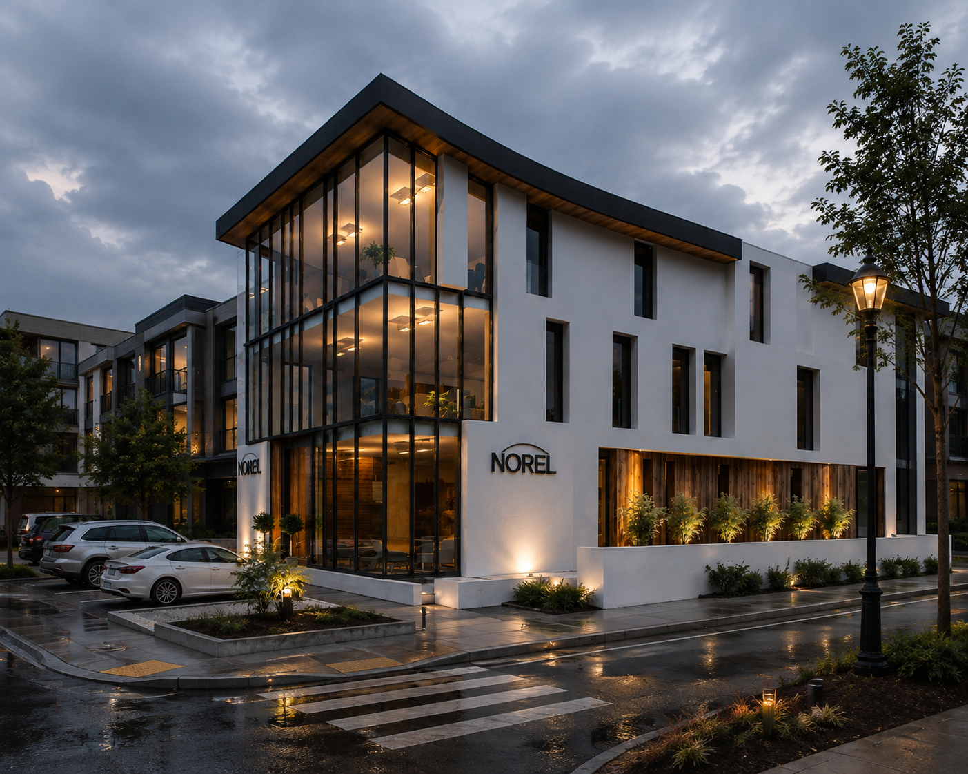 Modern multi-story building with large glass windows, illuminated from within, displaying the name 'Norel' on the exterior. The building is situated in a wet parking lot with several parked cars, small landscaped areas with plants and trees, and street lamps. The sky is cloudy, indicating dusk or early evening.