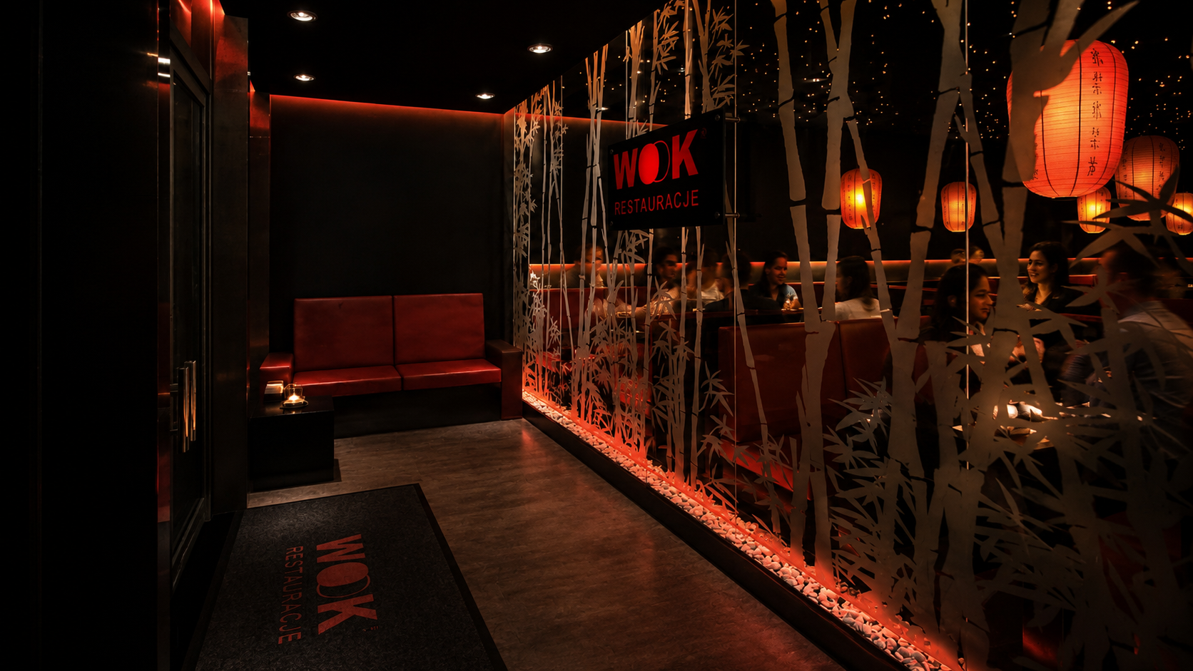 The image depicts the entrance of a dimly lit Japanese restaurant with a modern interior. There is a red cushioned bench on the left and a black mat with red text reading "WOK RESTAURACJE". A partition with bamboo and leaf designs, illuminated by orange lighting, separates the entrance from a dining area where several people are seated and enjoying their meals. Red paper lanterns hang from the ceiling, casting a warm glow over the space.