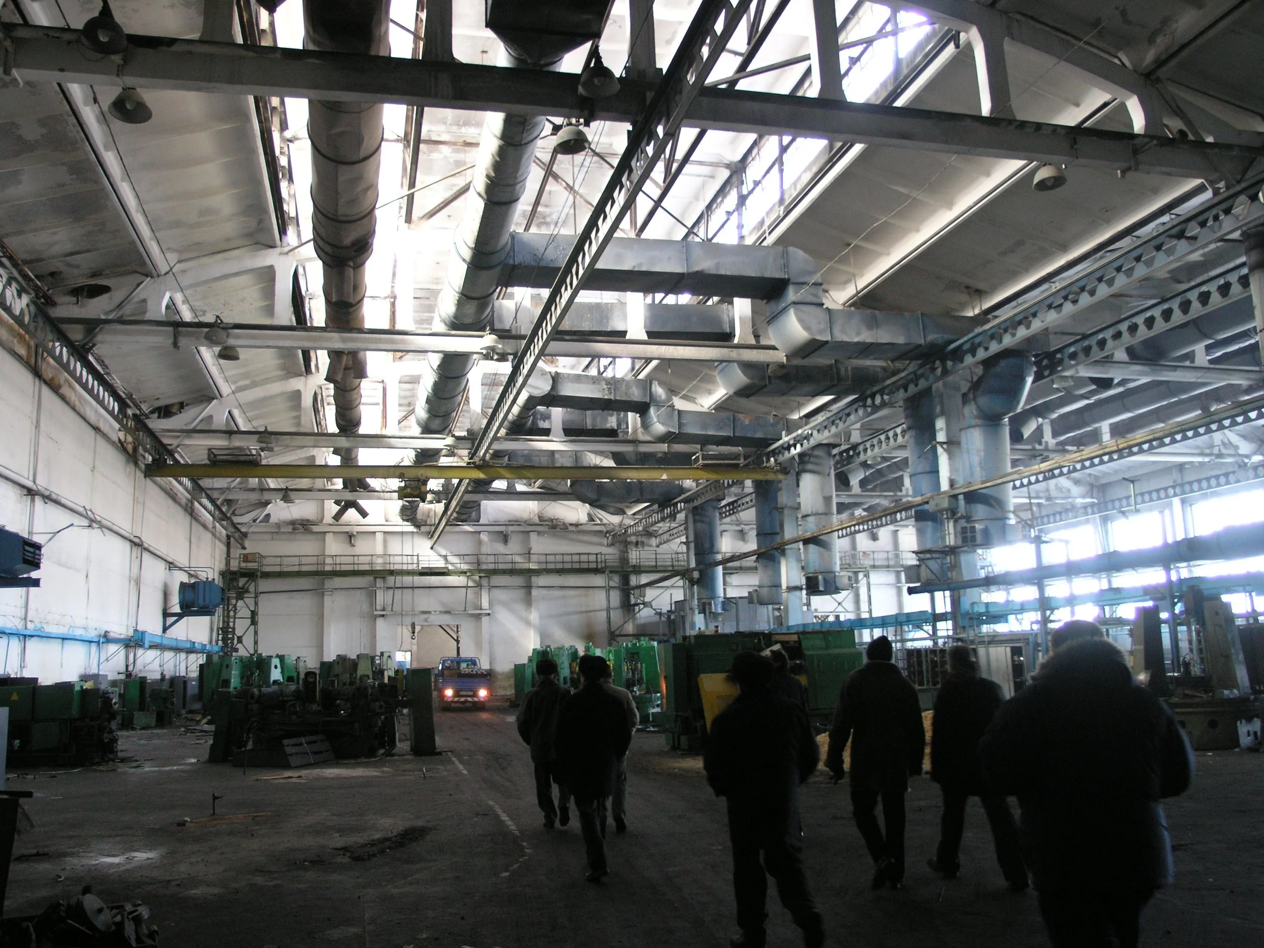 Interior of an industrial or factory building with high ceilings, large pipes, and metal beams. Several people are walking inside, and there are machinery and equipment along the sides.