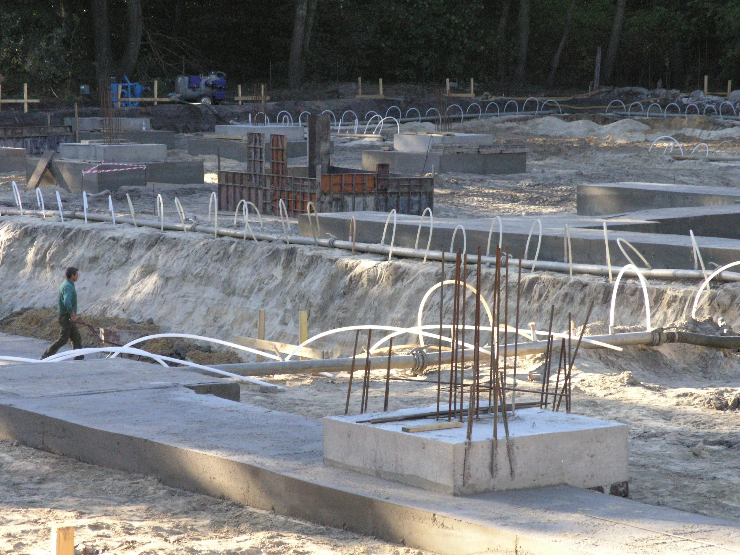 This is an image of a construction site with exposed concrete foundations and metal rebar. There are multiple curved white pipes and scattered construction materials. A person is walking in the foreground.