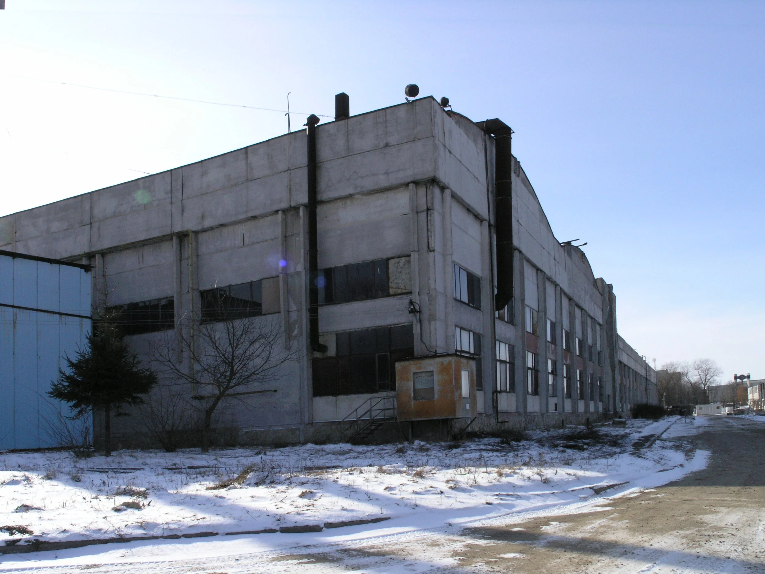 A large, multi-story concrete building with black pipes running down the side, and several small antennas on the roof, set against a clear blue sky. The ground in front is partially covered with snow, and there are some leafless trees and a street.