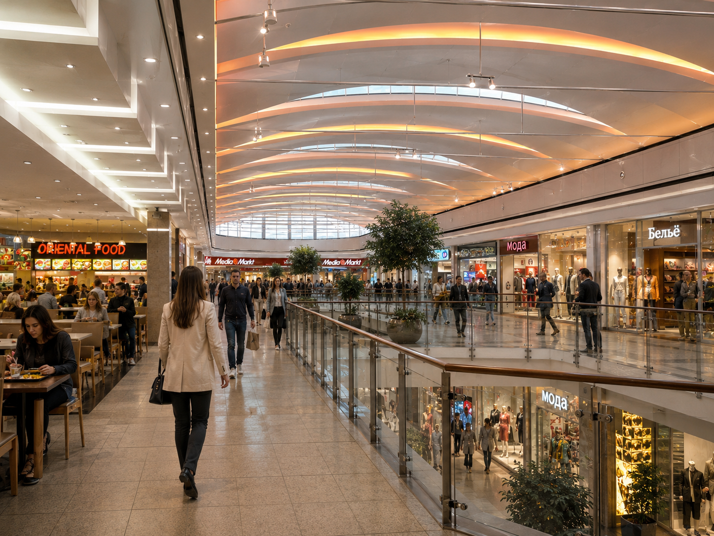 Interior of a shopping mall with multiple shoppers walking and sitting at a food court, including a woman in a beige coat walking towards the camera. The mall has bright lighting, trees in large planters, and storefronts including media market, fashion, and clothing stores visible on the upper level, with railings and glass barriers.