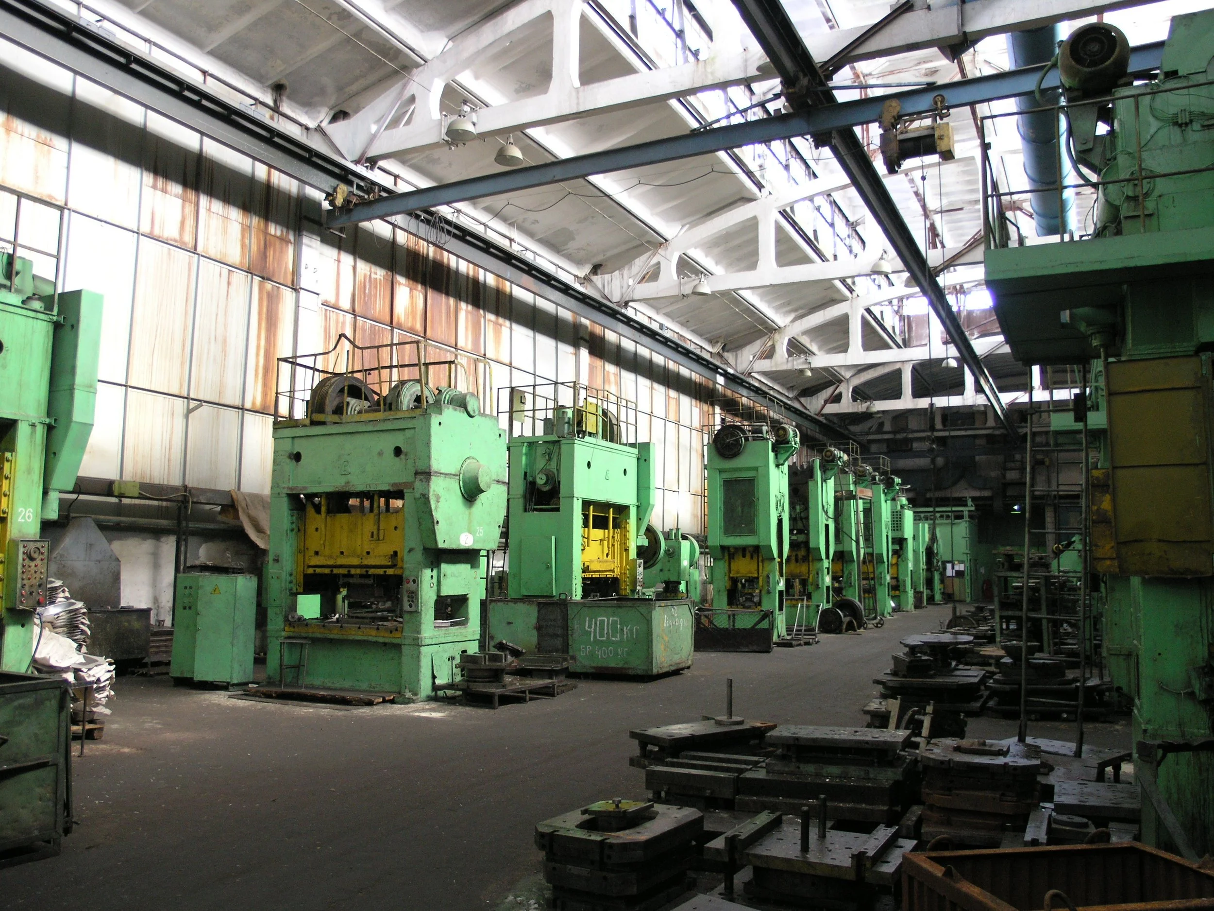 An industrial factory interior with green machinery, multiple tables, and a high ceiling with skylights and metal beams.