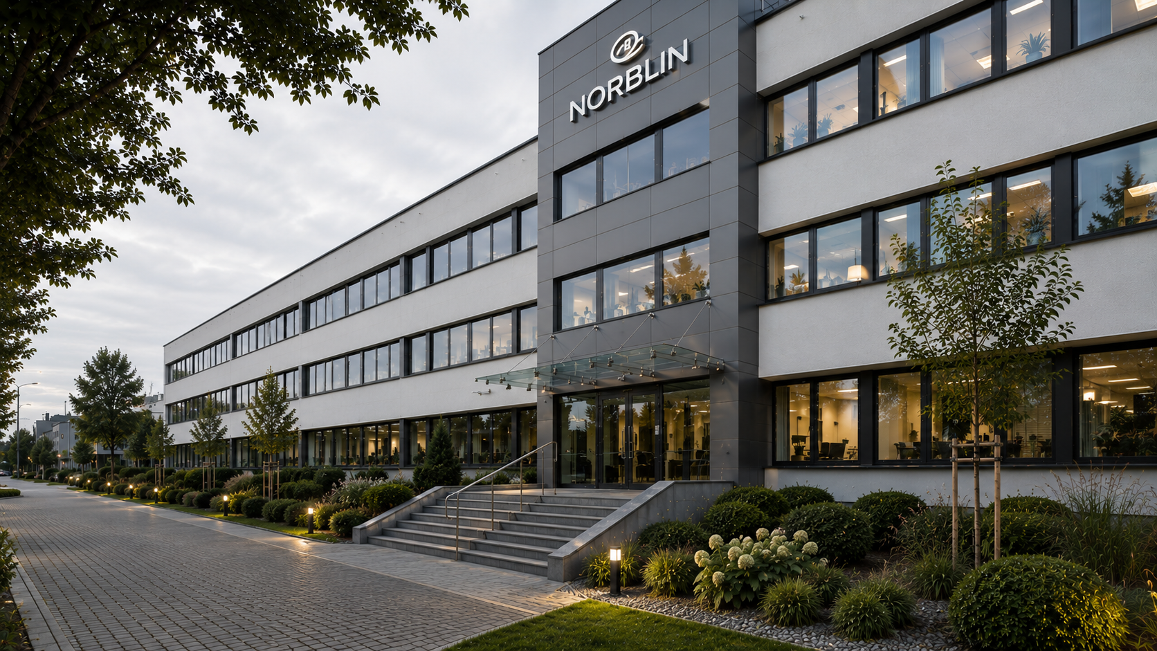 Modern office building with the 'Norblin' sign, glass entrance, landscaped surroundings, and multiple illuminated interior office spaces, during evening. Norblin, Warszawa