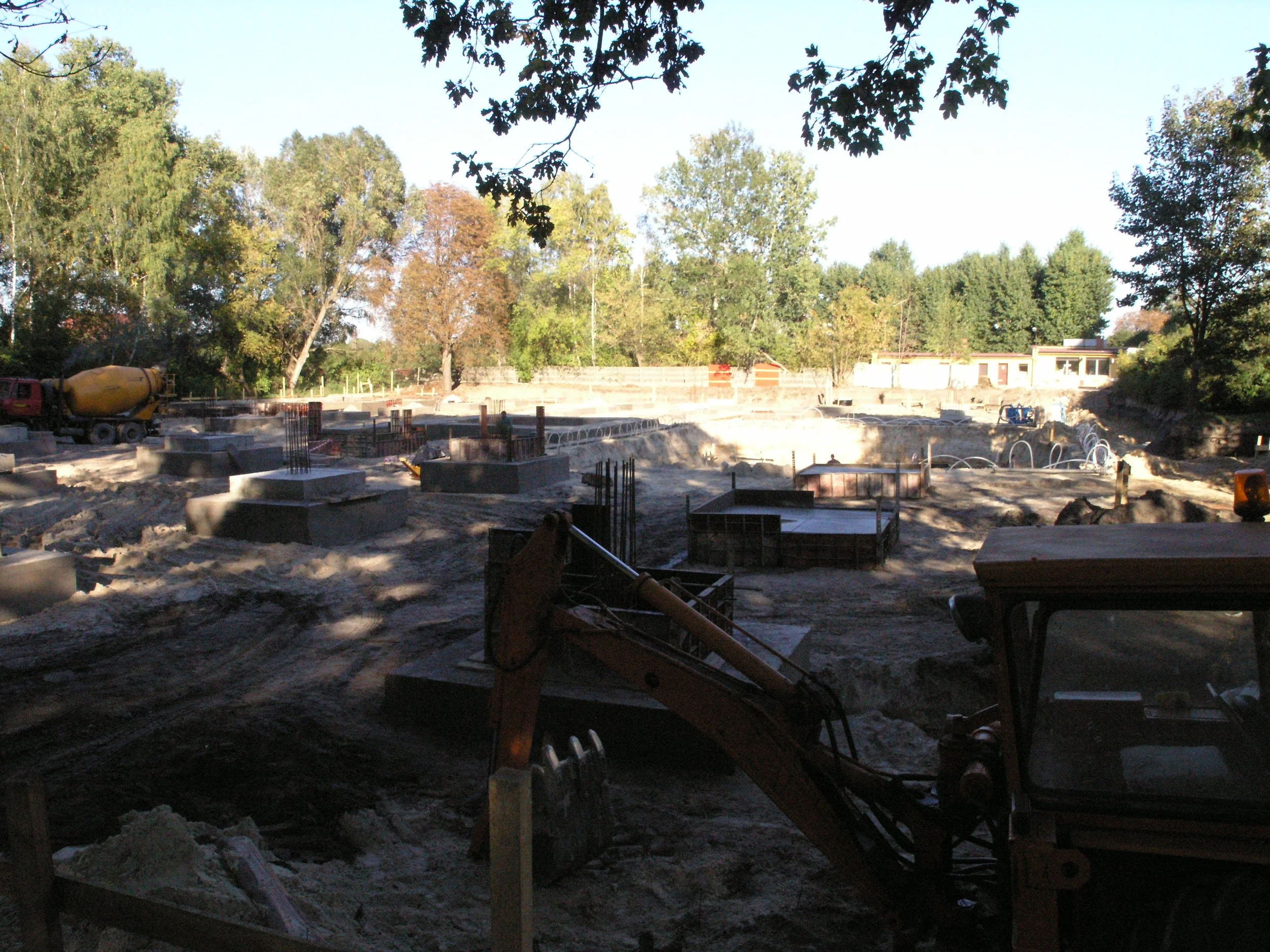 Construction site with foundation and structural elements, surrounded by trees in the background.