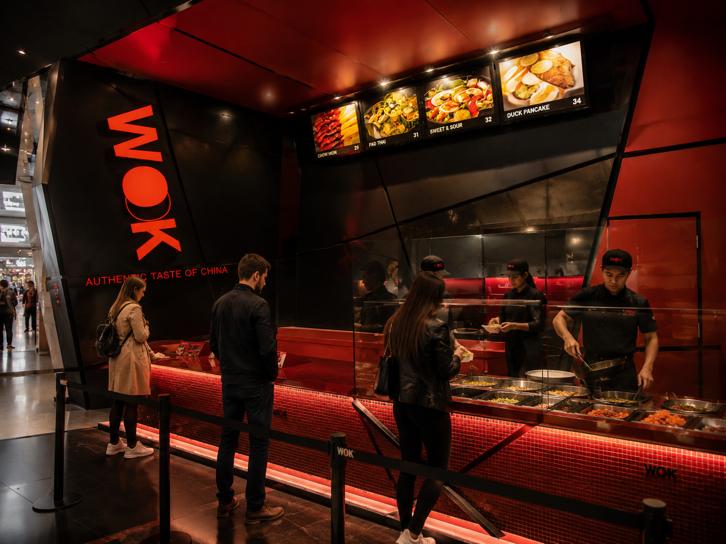 A food stall at a mall called WOK serving Chinese dishes with customers ordering food and staff preparing meals behind the counter. Wook