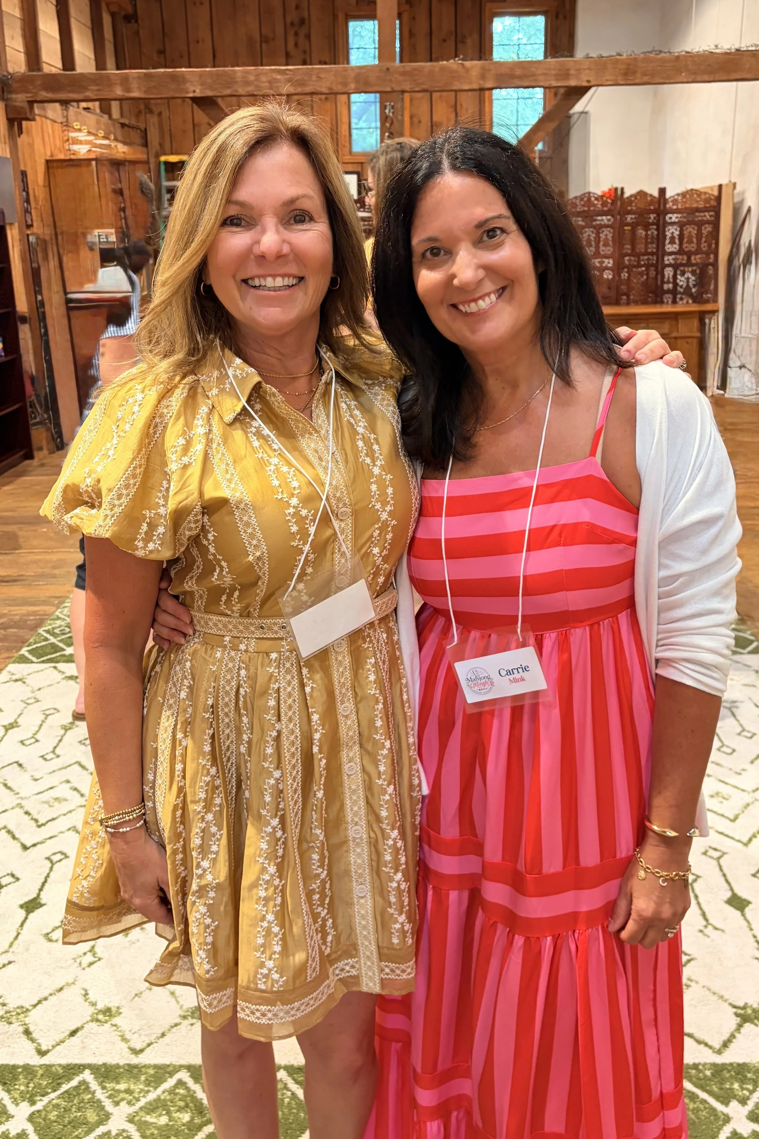 Two women smiling at a social gathering inside a rustic wooden building. One woman wears a yellow dress with white embroidery, the other wears a pink and red striped dress with a white cardigan. Both have name tags and are wearing earphones.