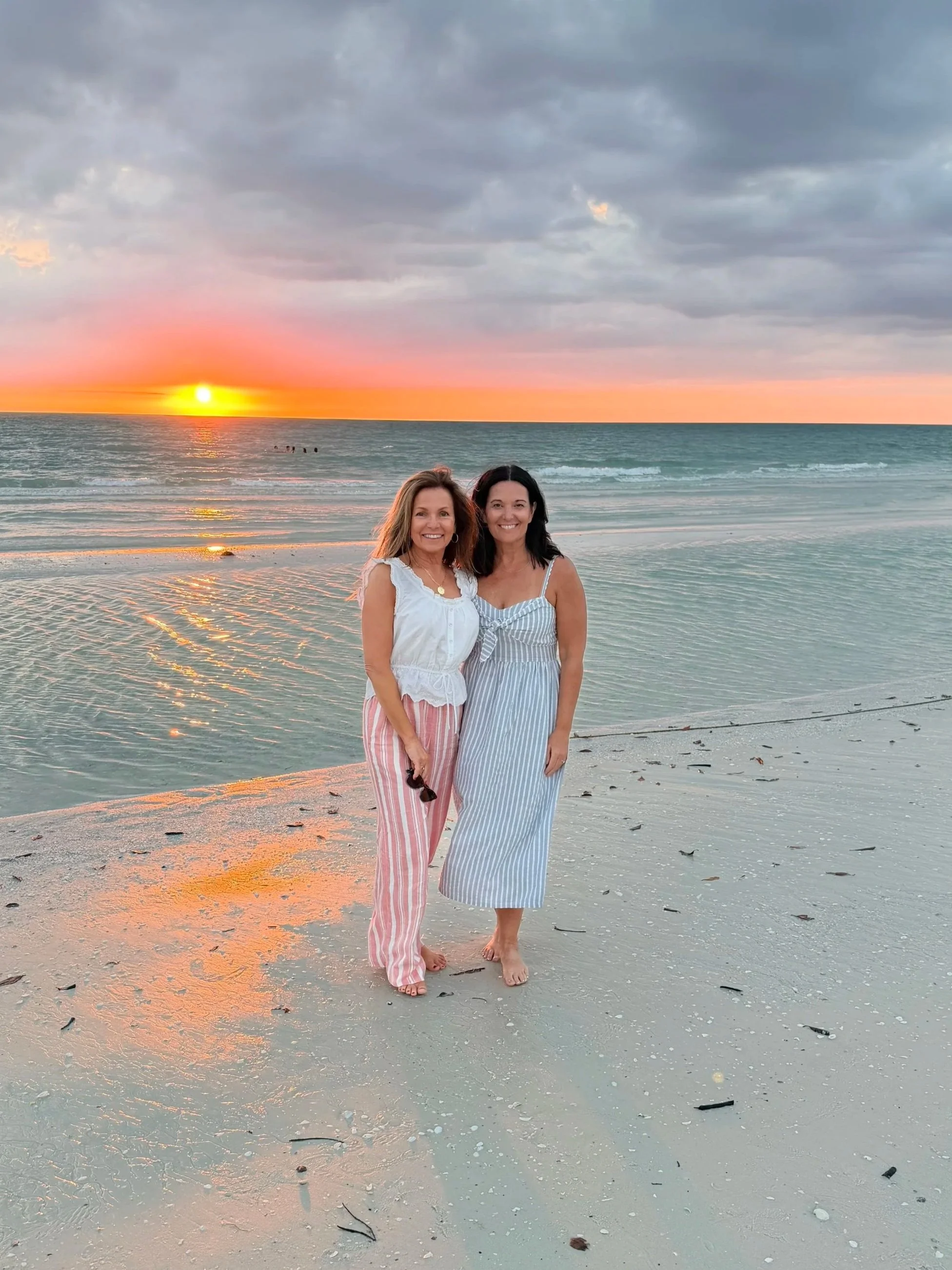 Two women standing on a beach at sunset, smiling, with dark clouds overhead and calm ocean waves in the background.