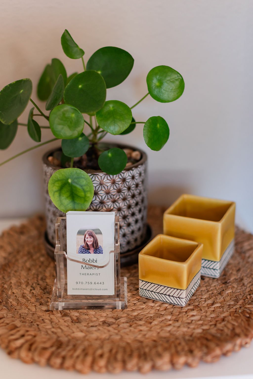 A potted plant with round green leaves, a business card holder with a photo and contact information for Bobbi Maier, a therapist, and two yellow and white striped square containers, all arranged on a round woven mat.