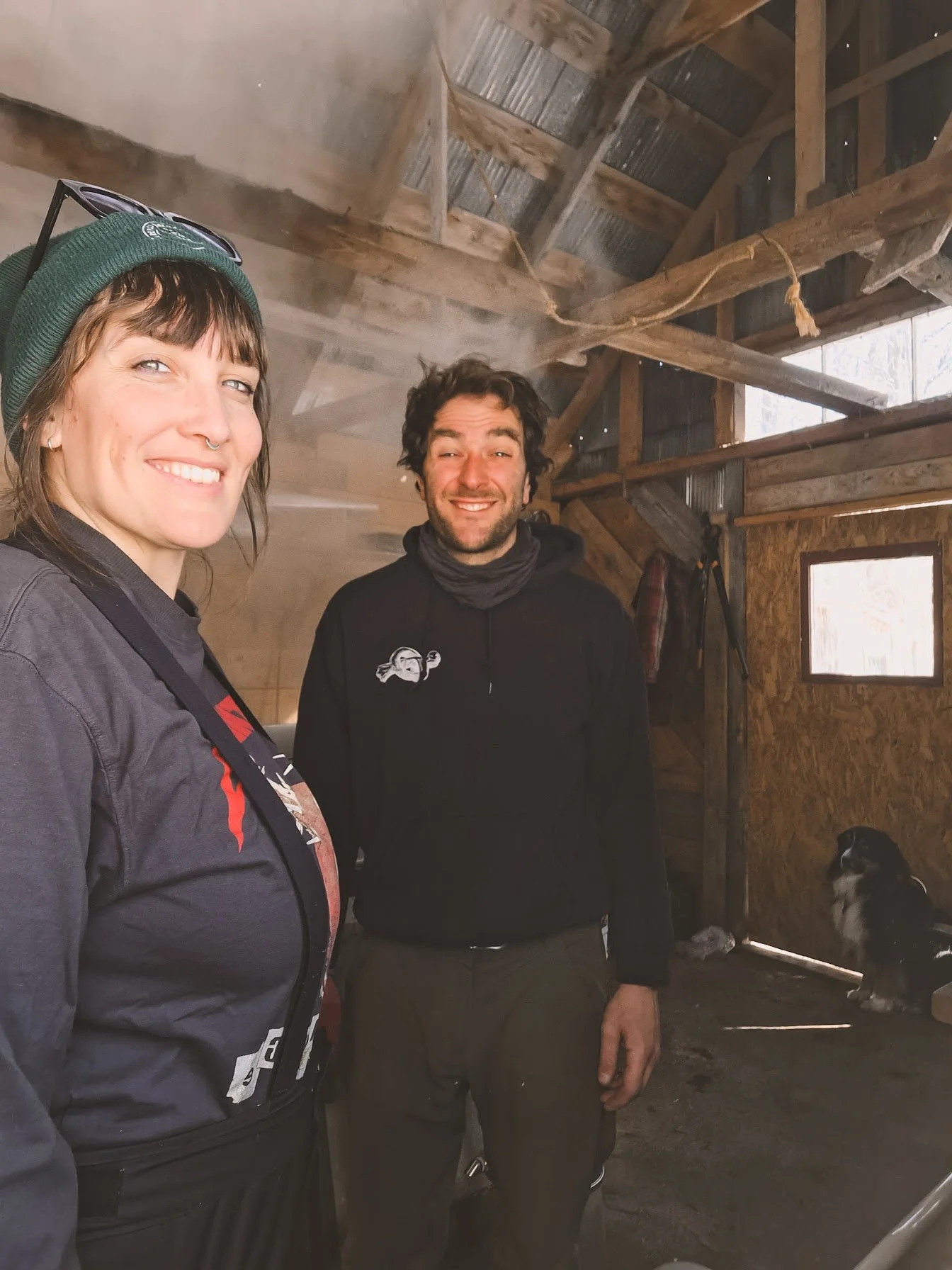 Deux personnes posent pour une photo dans une ferme en bois avec de la fumée. Un chien de berger, probablement un husky, est visible dans le coin inférieur droit.