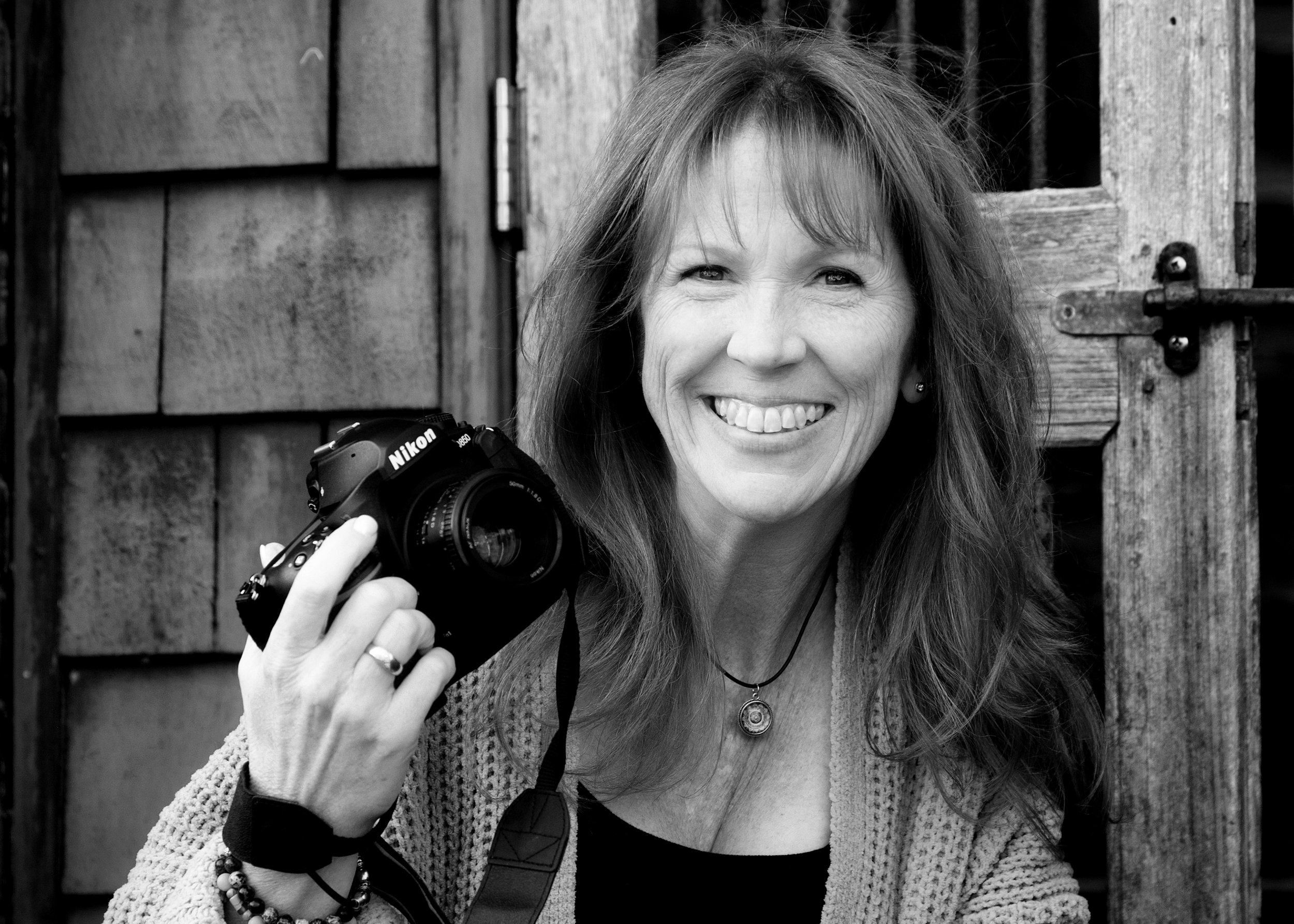 Melody McBride, photographer and Director, smiling, holding a Nikon camera, standing outdoors in front of a wooden structure.