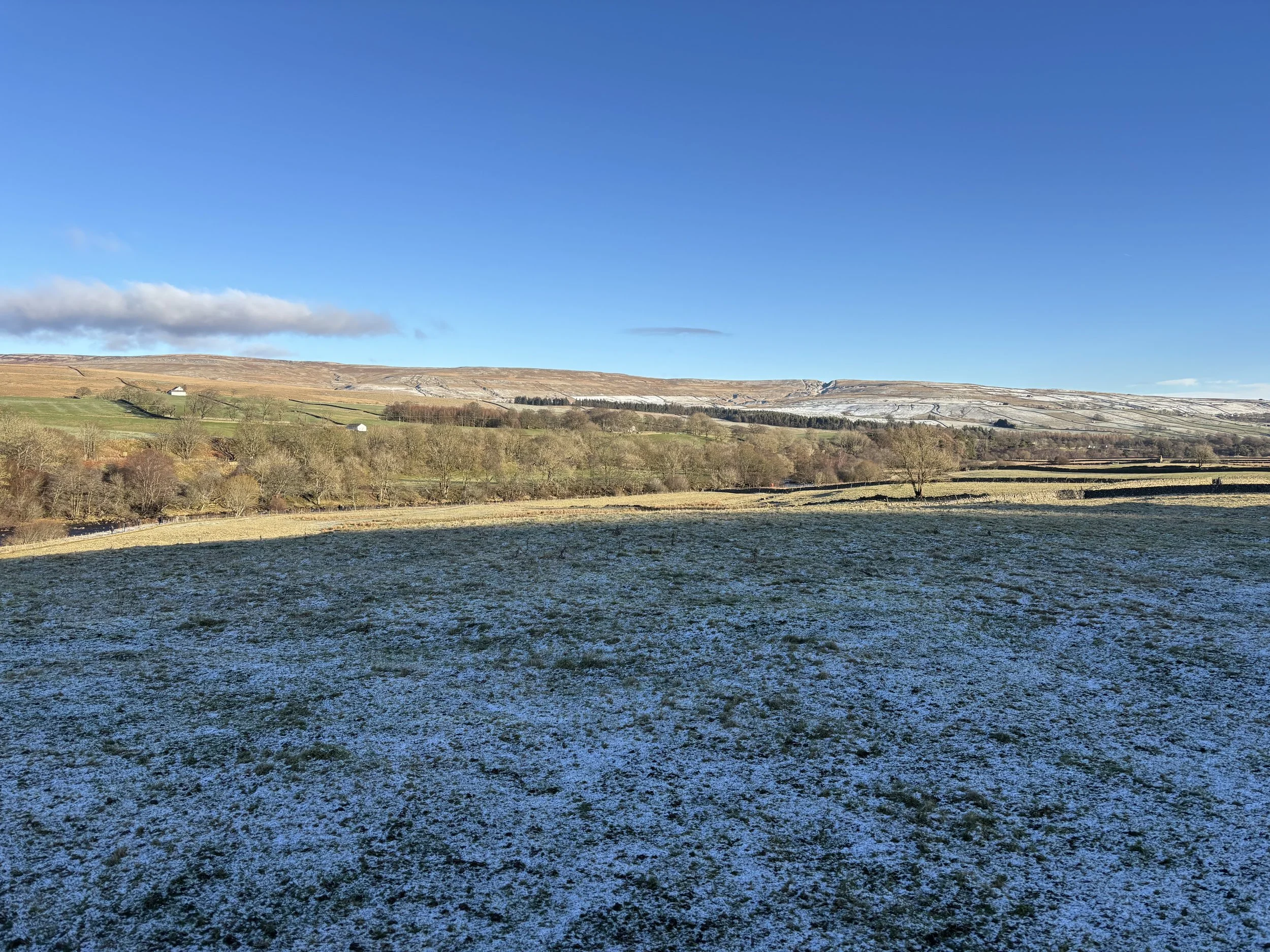 Snow-dusted countryside with rolling hills, patchwork fields, scattered trees, and farm buildings beneath a clear blue sky
