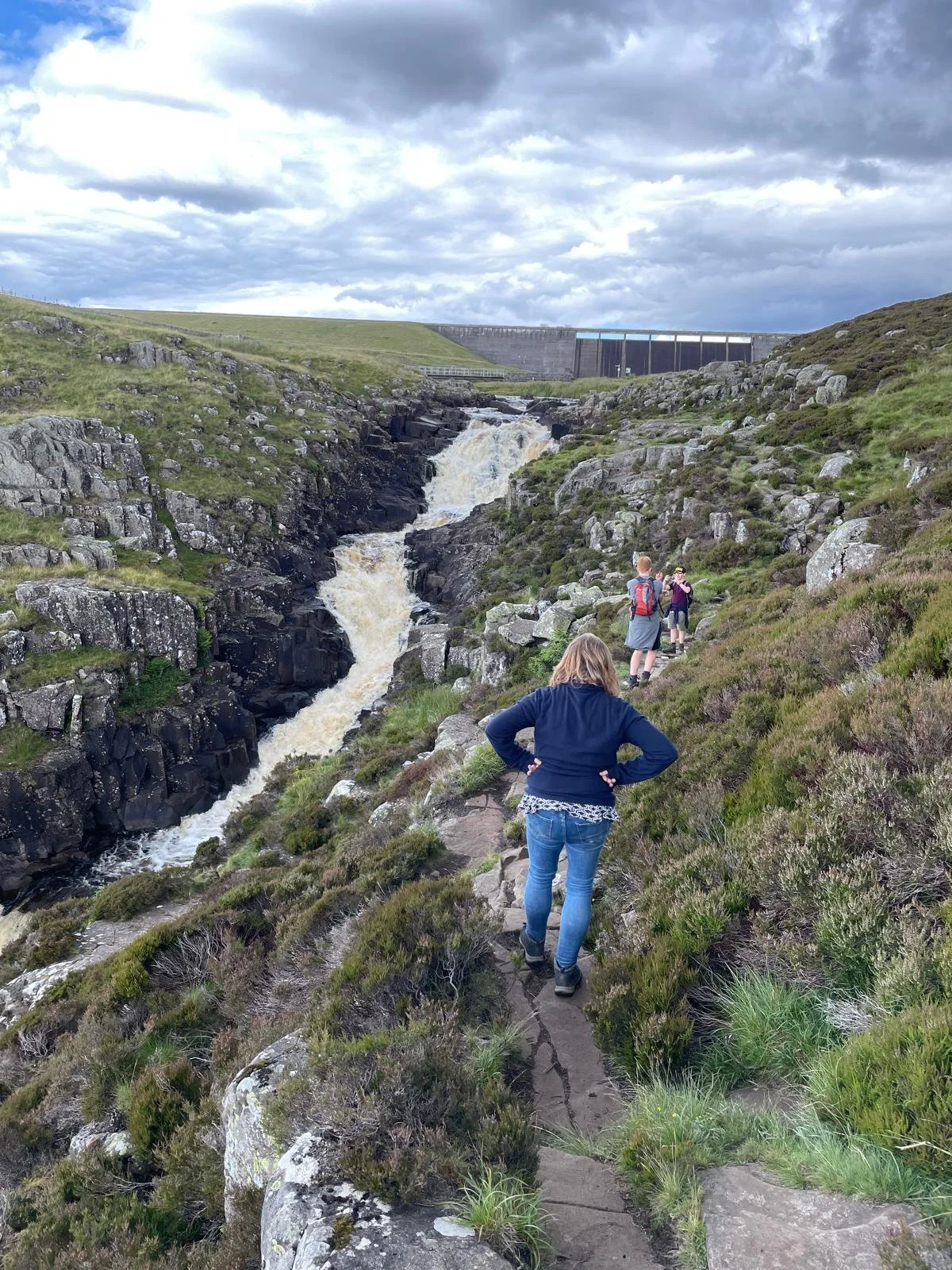 Hikers beside a rocky mountain waterfall below a concrete dam under cloudy skies