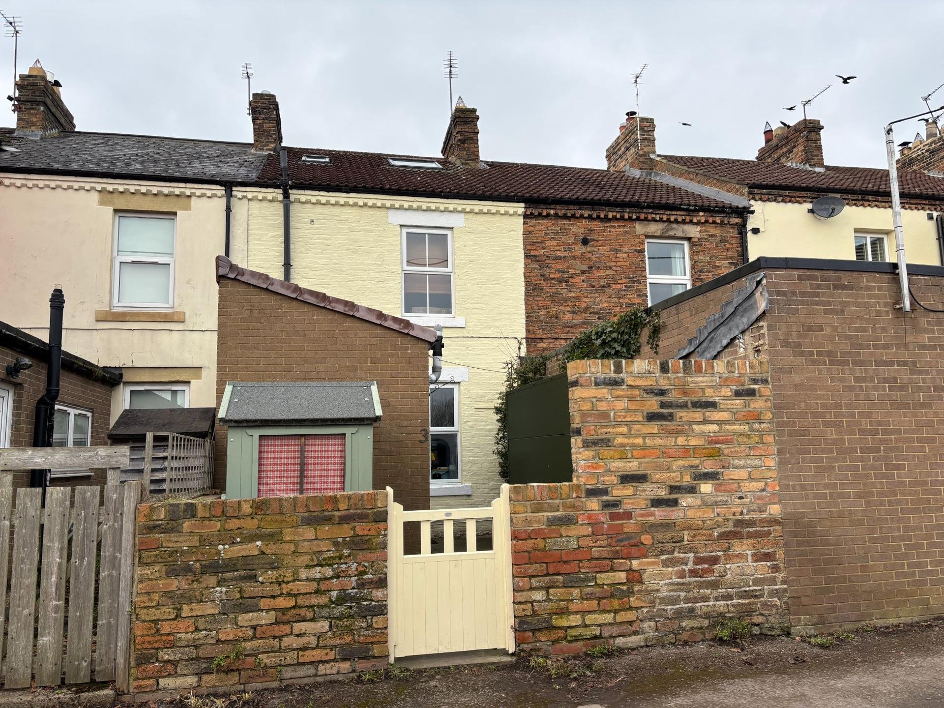 Row of attached brick terraced houses with tiled roofs, chimneys, backyard walls, and a small cream-colored gate