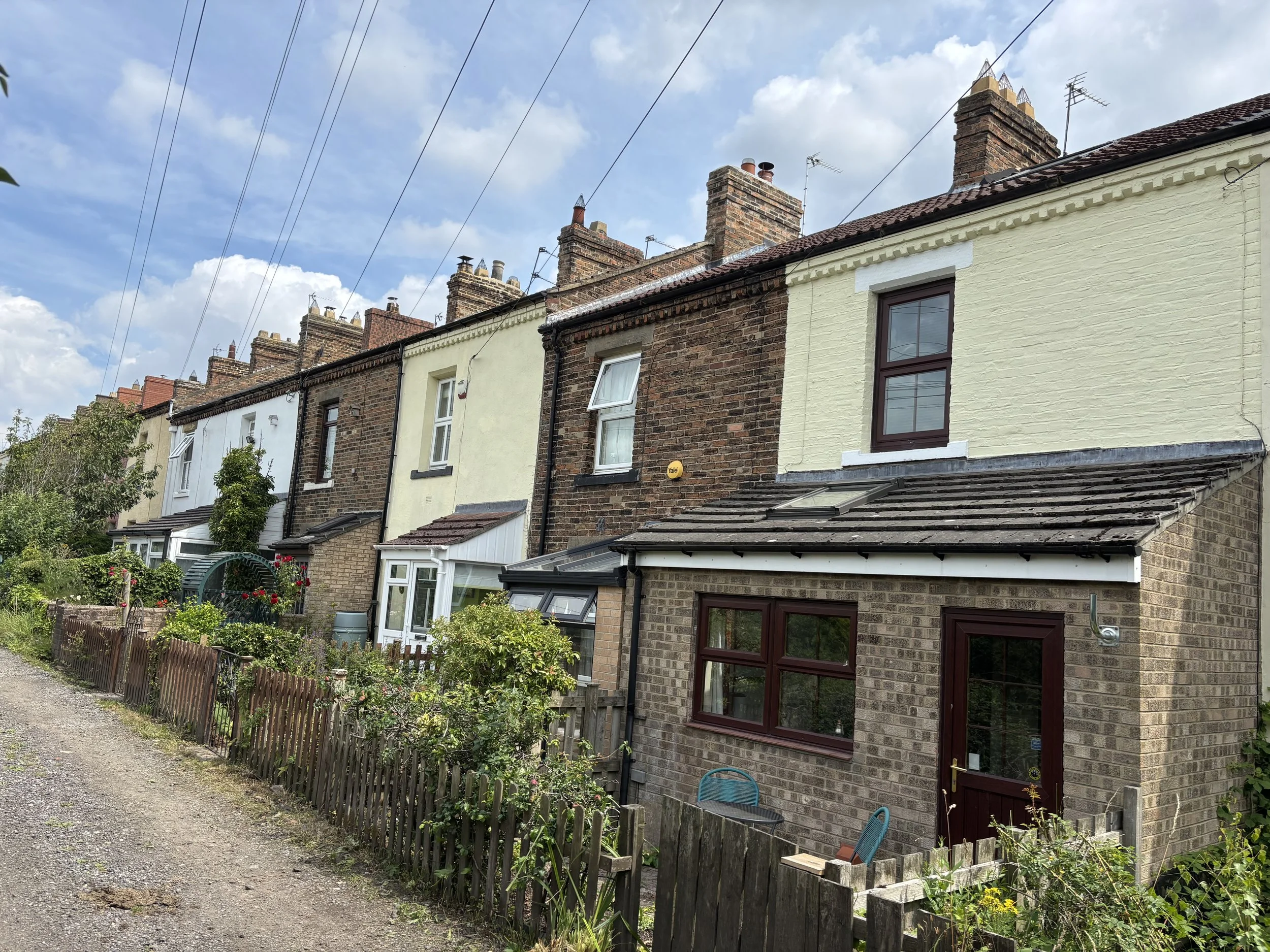 Row of brick terraced houses with tall chimneys, gardens, fences, and overhead utility wires under a partly cloudy sky.