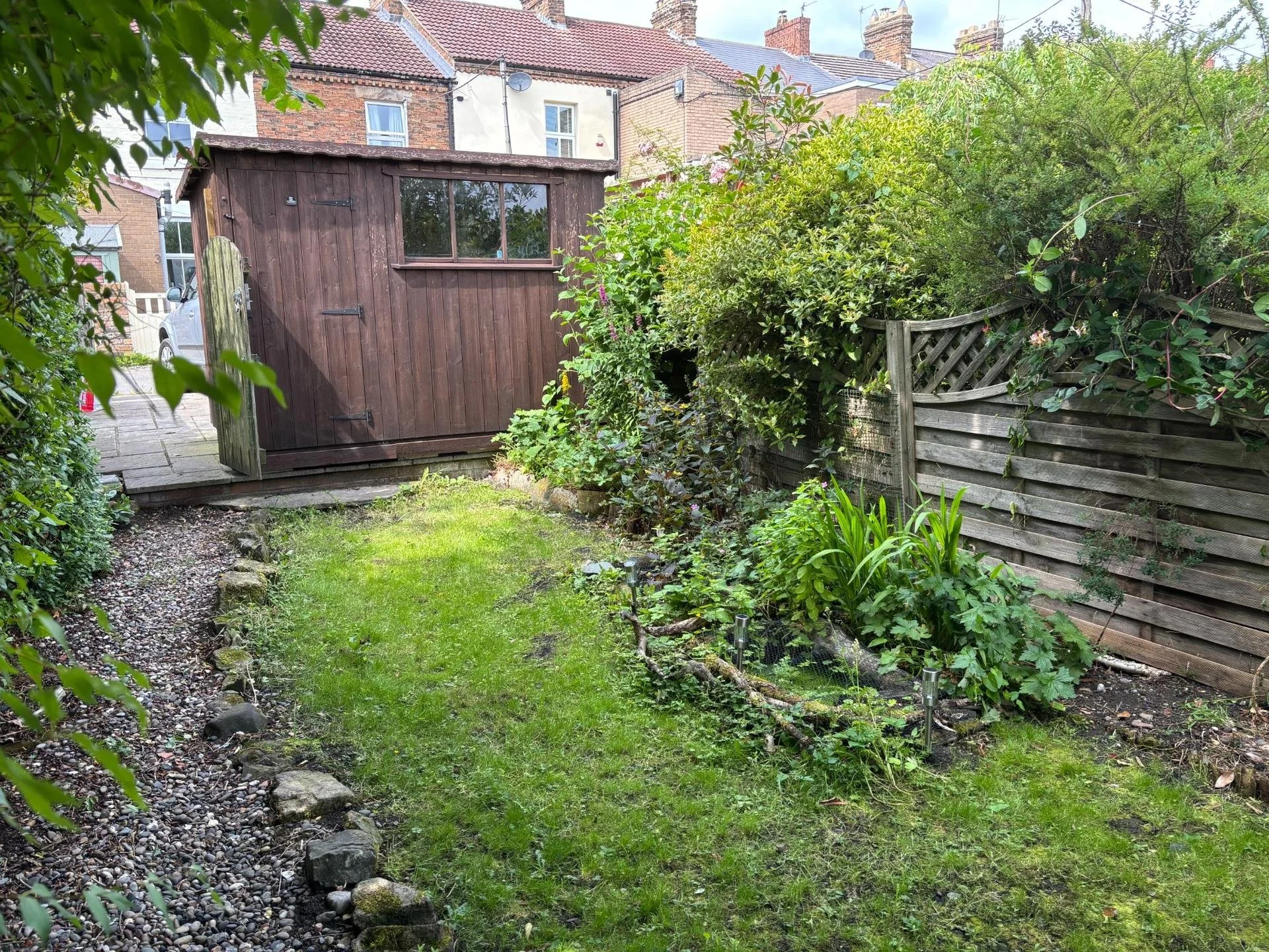 Lush backyard garden with a dark wooden shed, raised planting beds, gravel path, and brick row houses in the background