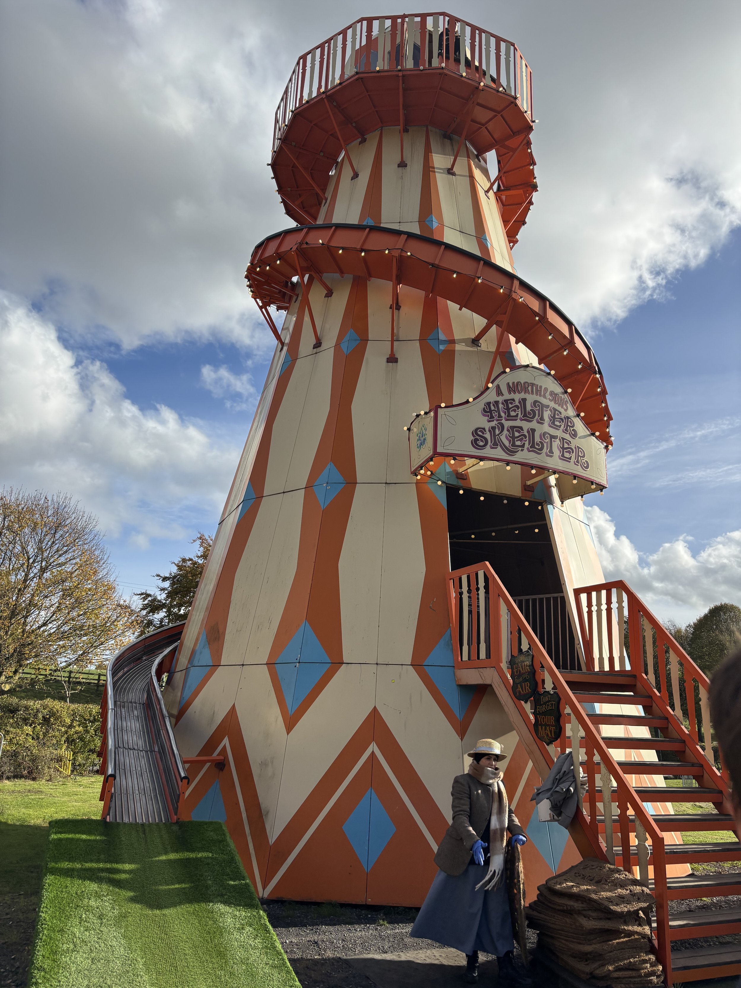 Colorful helter-skelter fairground slide with string lights, viewed from below against a partly cloudy sky