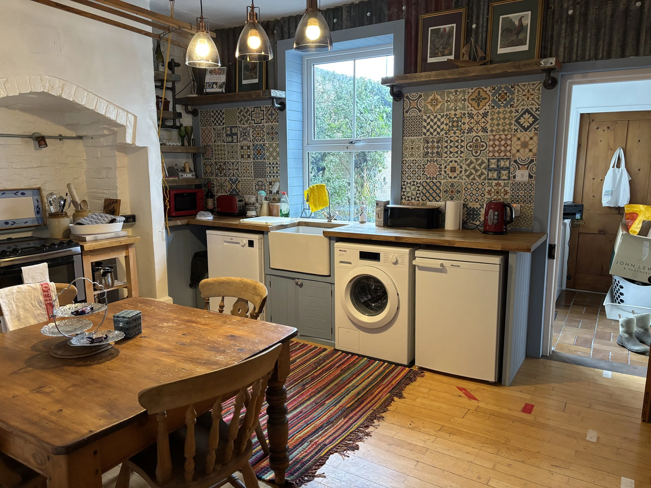 Rustic country kitchen with patterned tile backsplash, farmhouse dining table, wood stove, pendant lights, and built-in laundry appliances