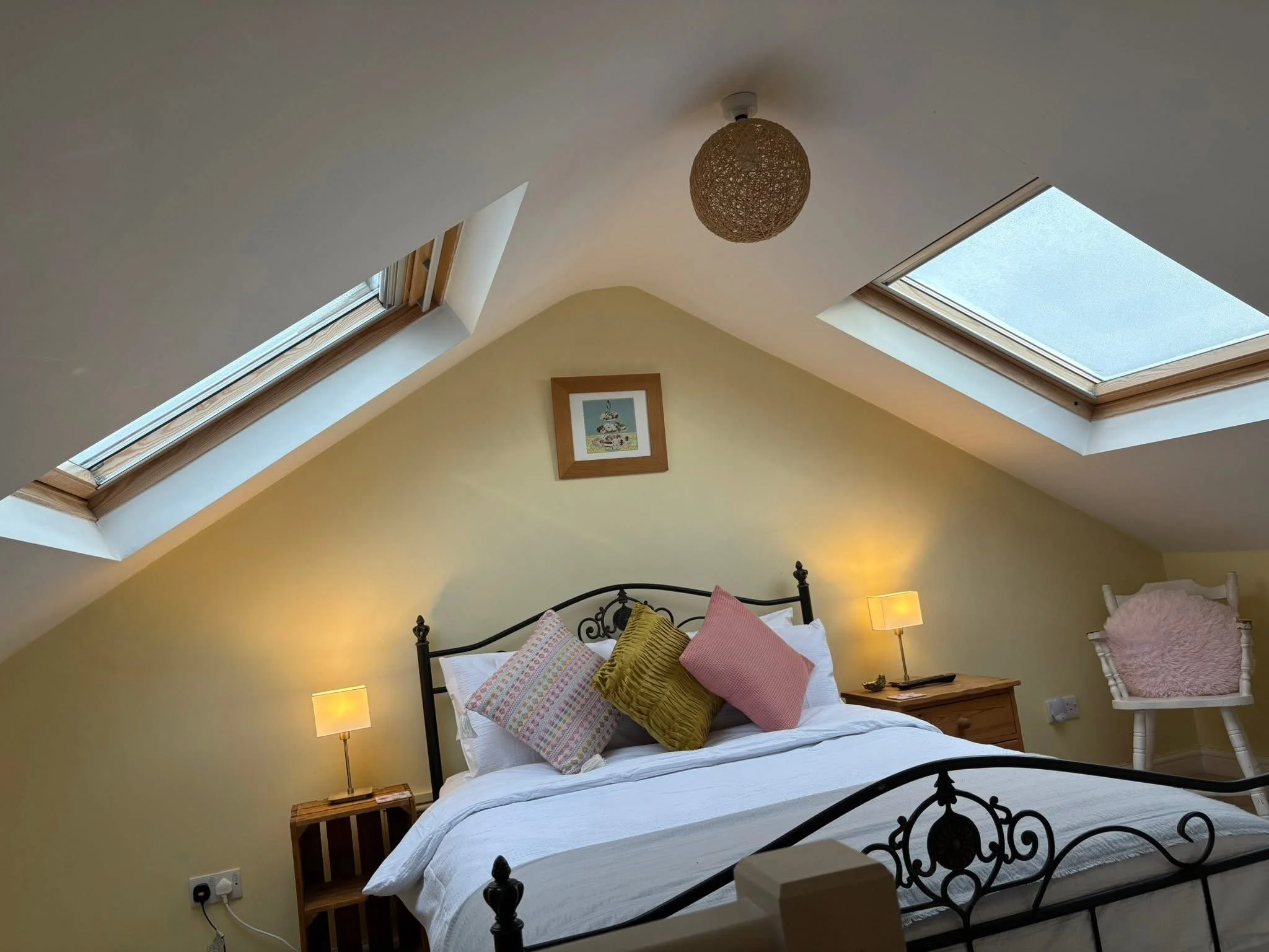Cozy attic bedroom with a wrought-iron bed, colorful pillows, skylights, bedside lamps, and a rocking chair.