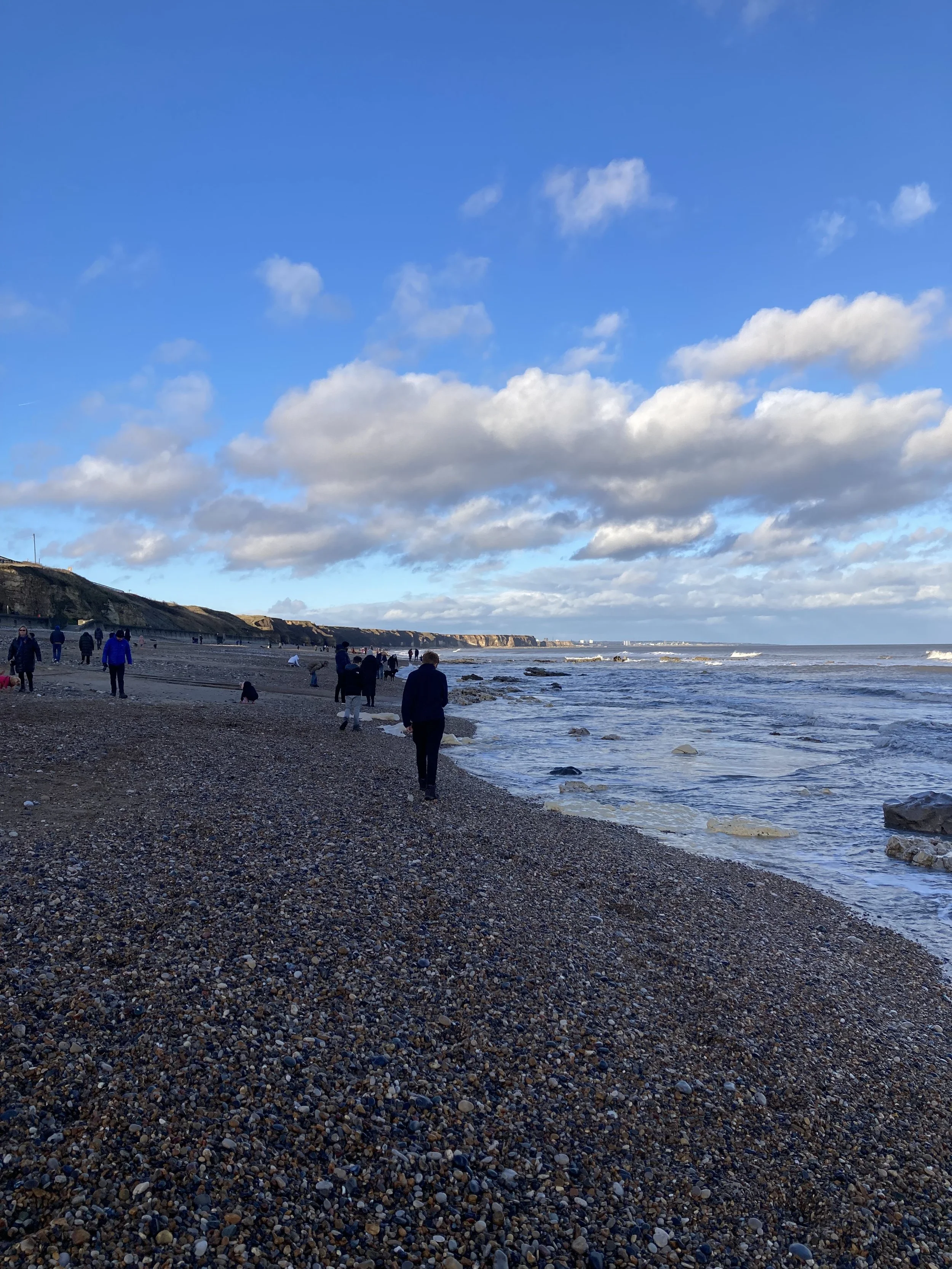 People walking along a rocky pebble beach beside choppy ocean waves and coastal cliffs under a blue, cloudy sky