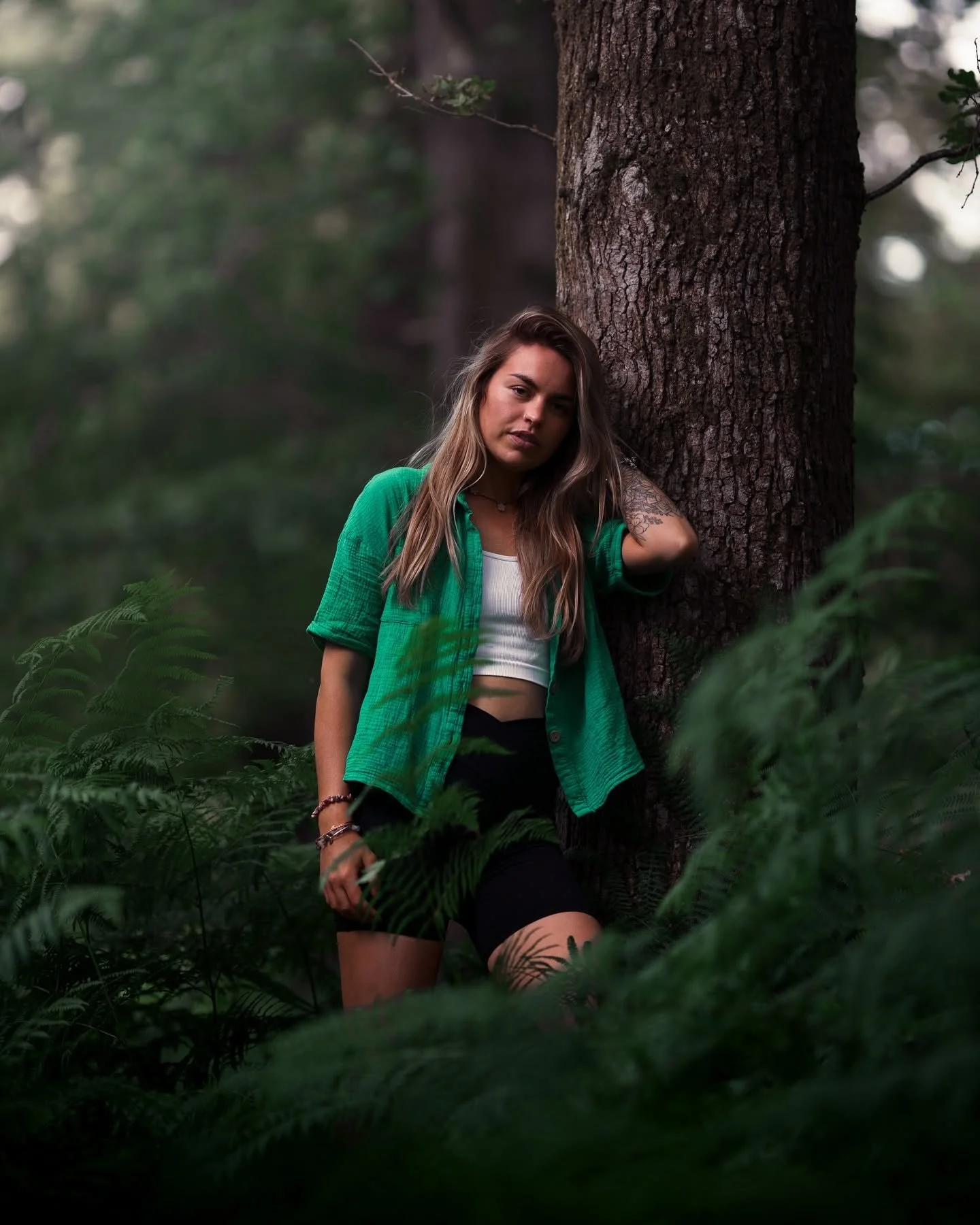 A young woman with long hair leaning against a tree in a forest surrounded by green ferns. She's wearing a green shirt over a white top and black shorts.