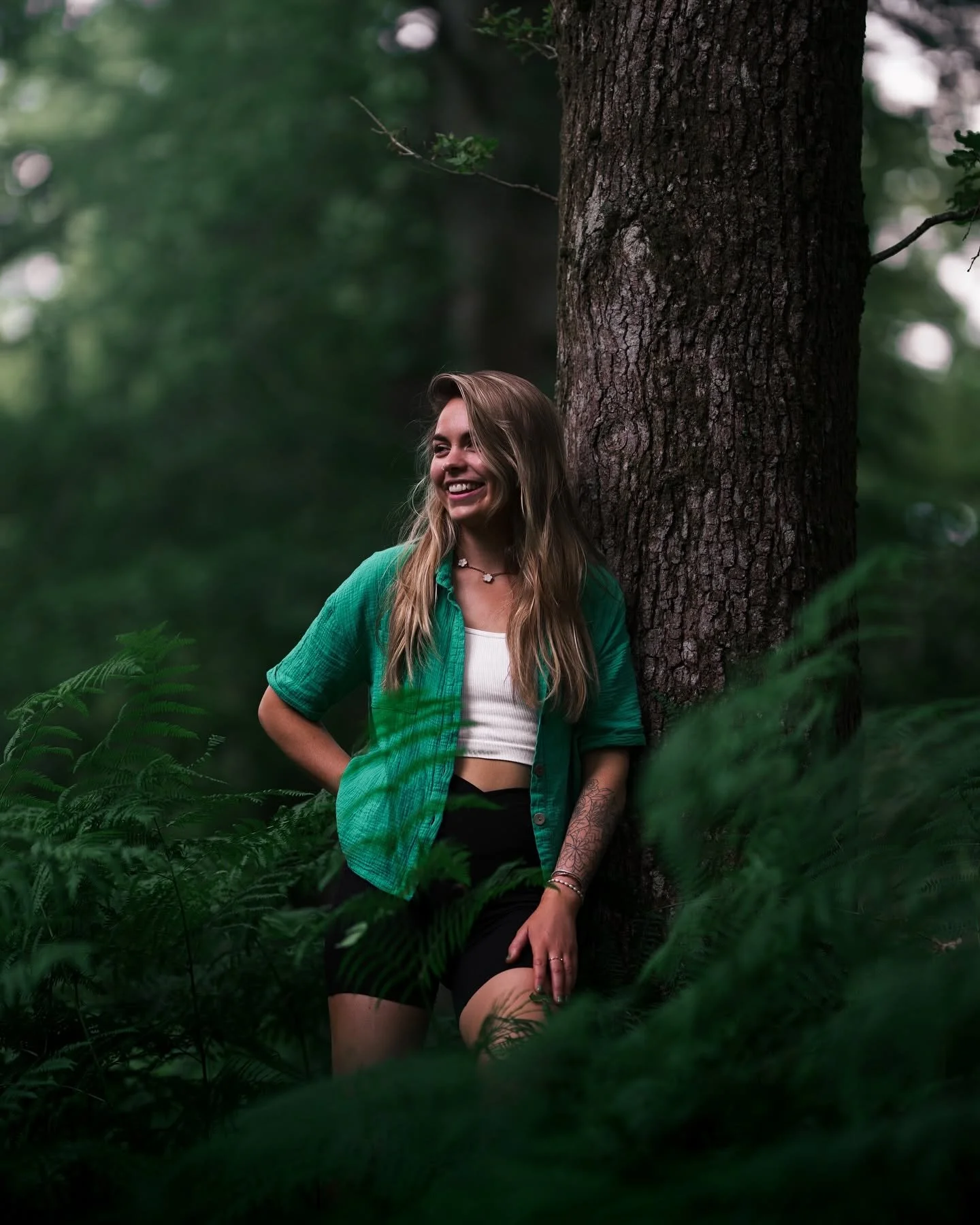 Young woman with long blonde hair, smiling, leaning against a tree trunk in a dense forest, wearing a green shirt over a white crop top, black shorts, and jewelry, surrounded by green foliage.