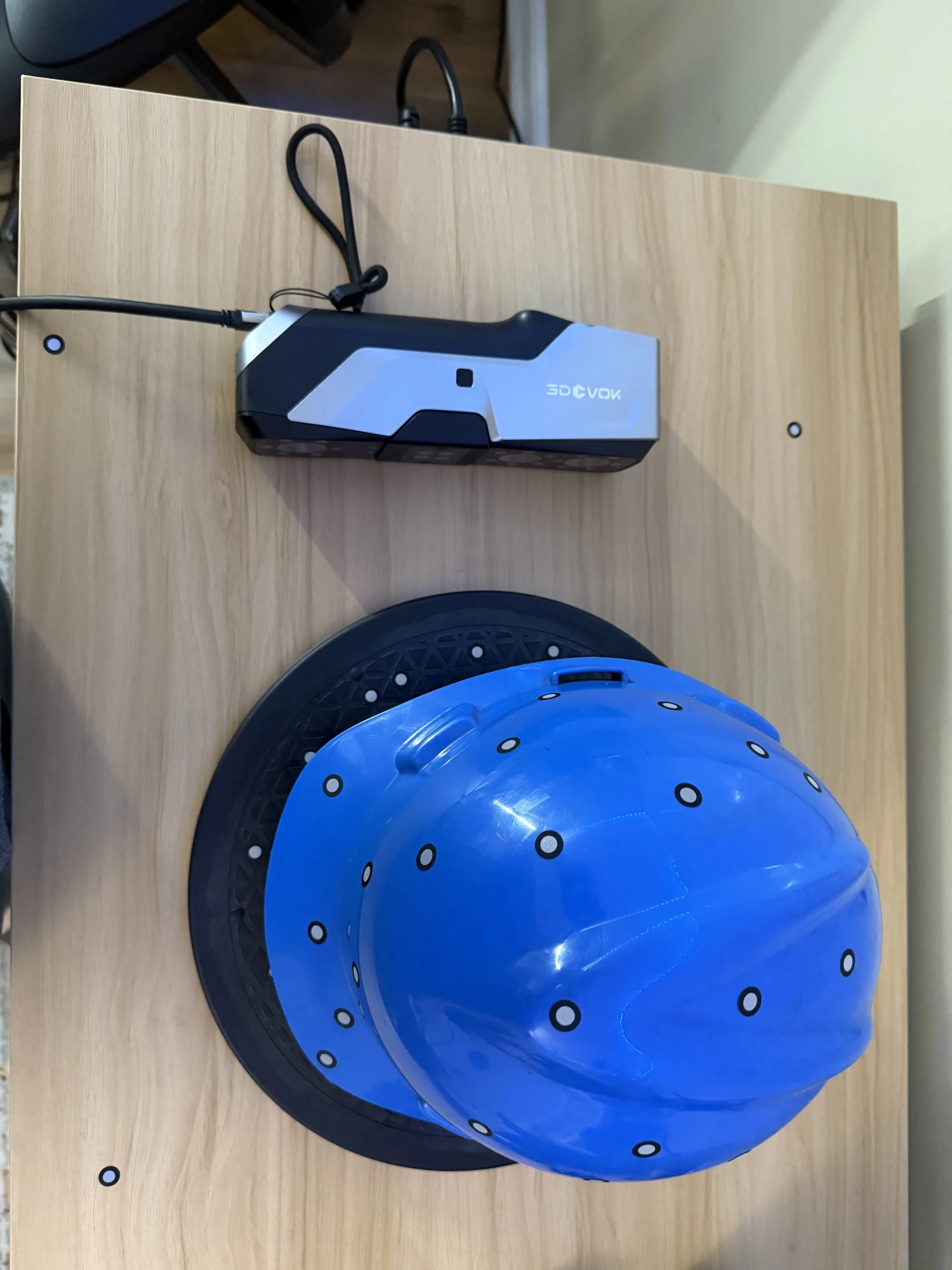 A blue ball with black and white dotted pattern placed on a black circular stand on a wooden surface with a beige wall in the background.