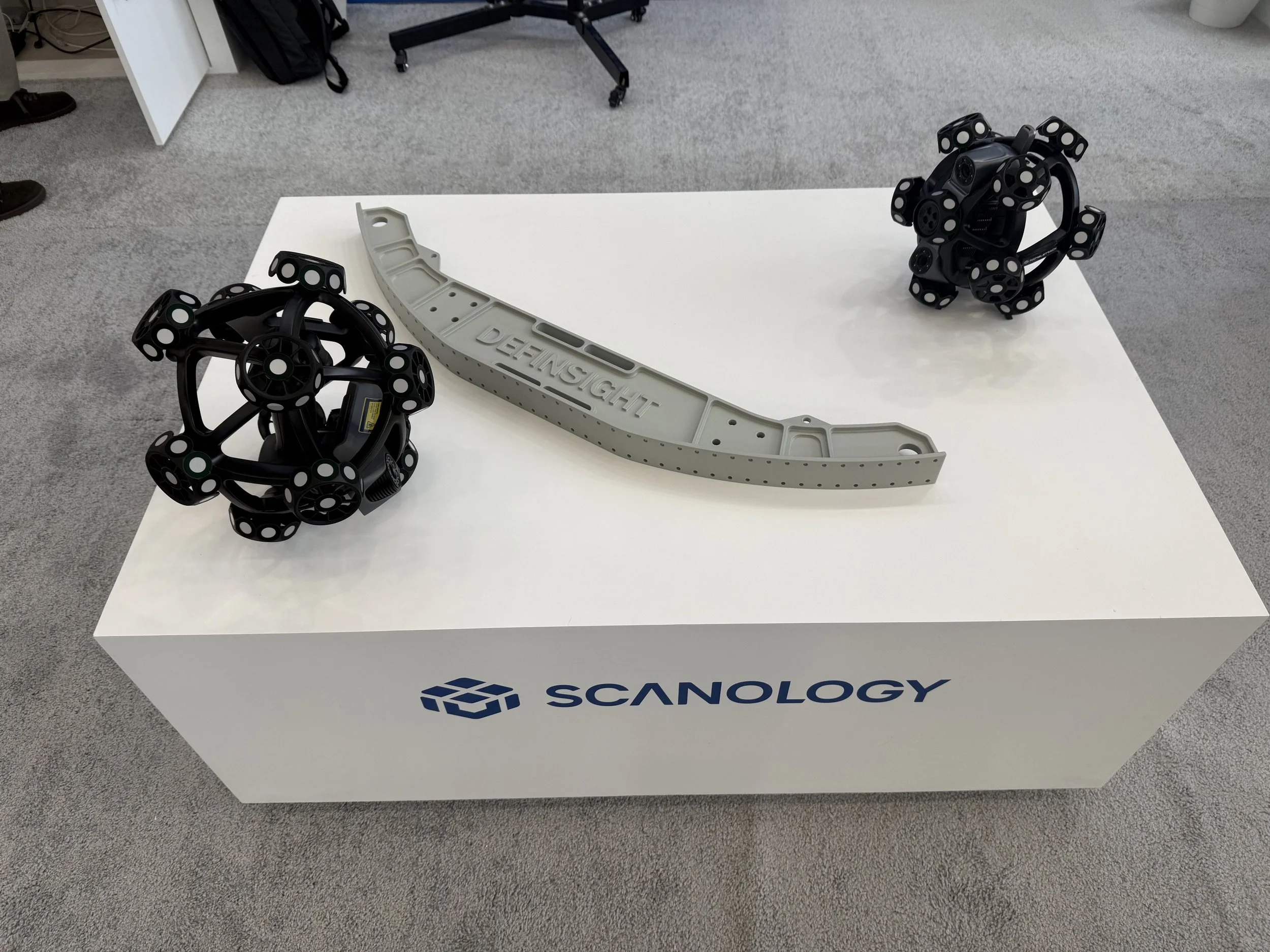 A white table with the Scanology logo, displaying two black spherical robotic structures with multiple circular openings and a gray elongated part with the word "DEIN SIGHT" on it, in a room with gray carpet and office furniture.