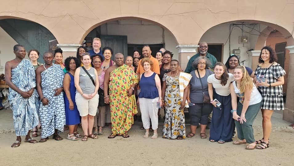 Group of diverse people standing together in front of an arched building, some wearing traditional African clothing, smiling for the photo.