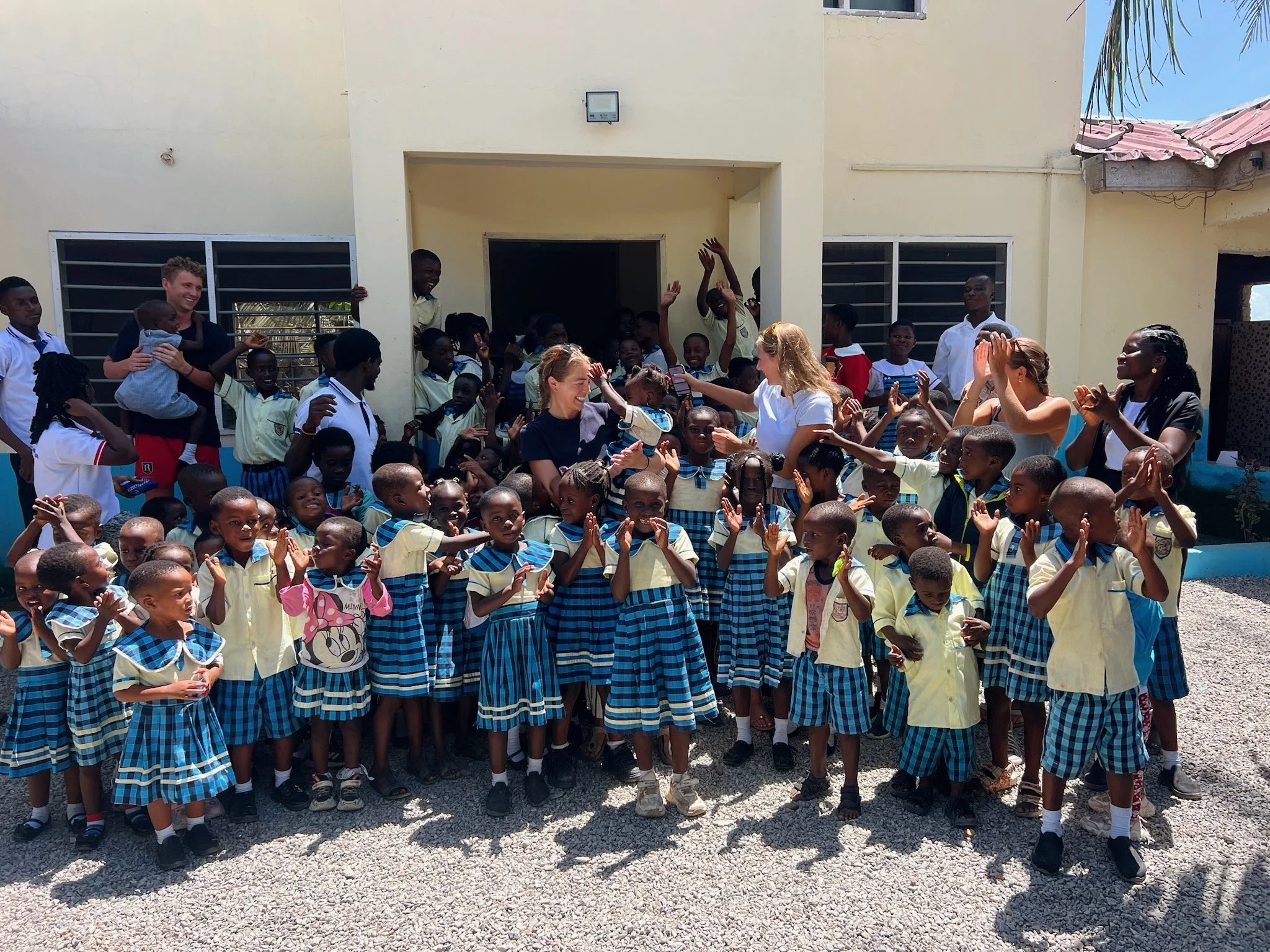 Group of children and adults gathered outside a building, smiling and waving, with some children wearing school uniforms and others in casual clothing.