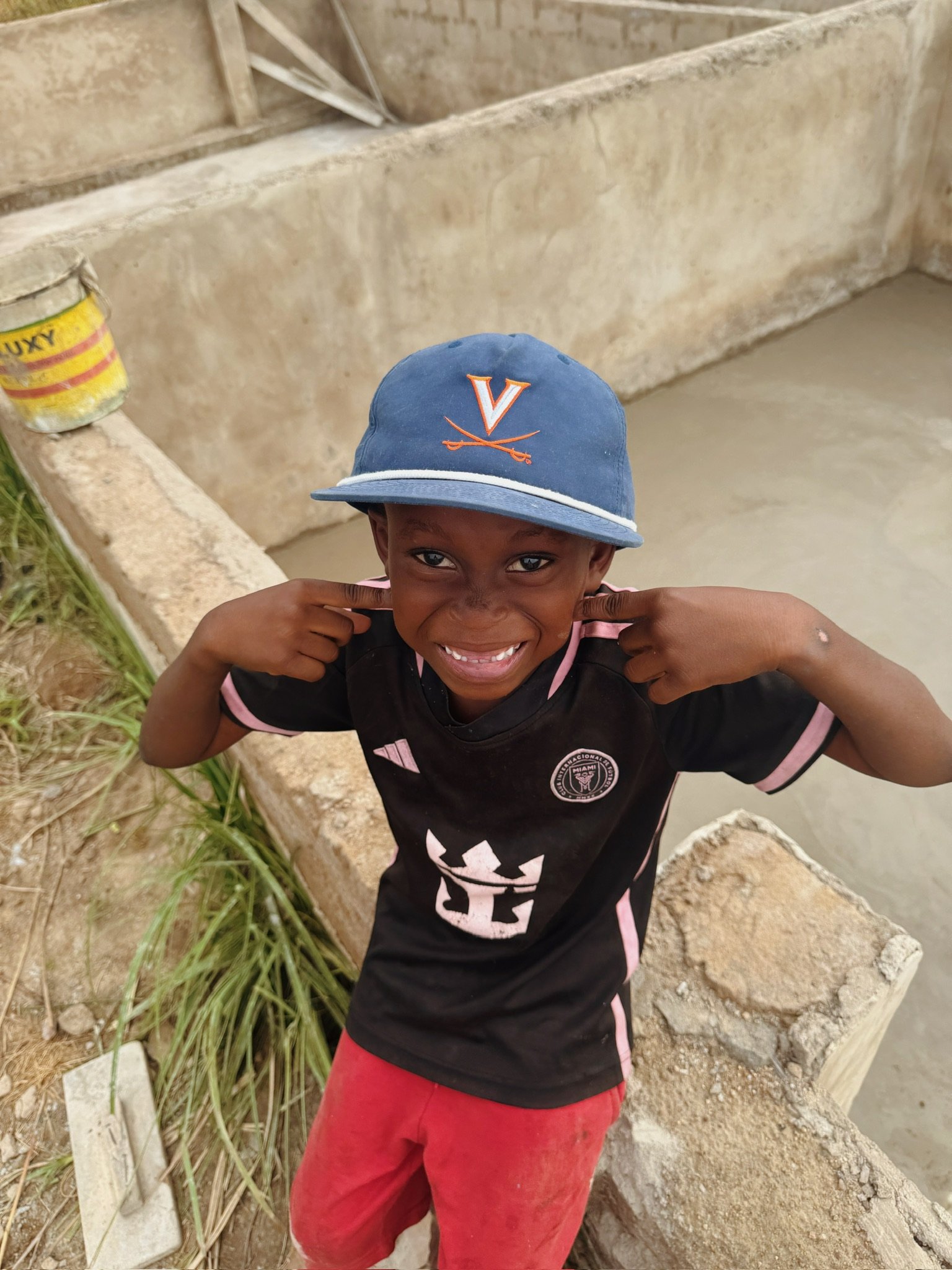 A young boy is smiling and pointing to his ears while standing near a concrete structure with water behind him. He is wearing a blue baseball cap with a Virginia Cavaliers logo, a black t-shirt with pink trim, and red pants.