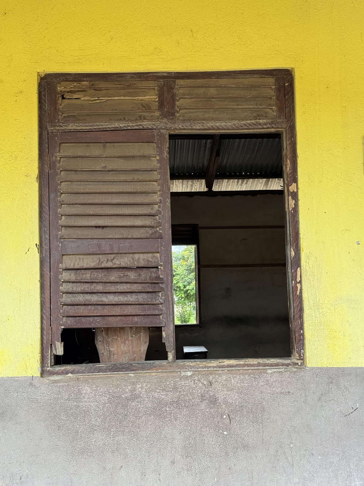 A yellow wall with an open wooden window frame, revealing an unfinished room with a corrugated metal roof and a window showing greenery outside.