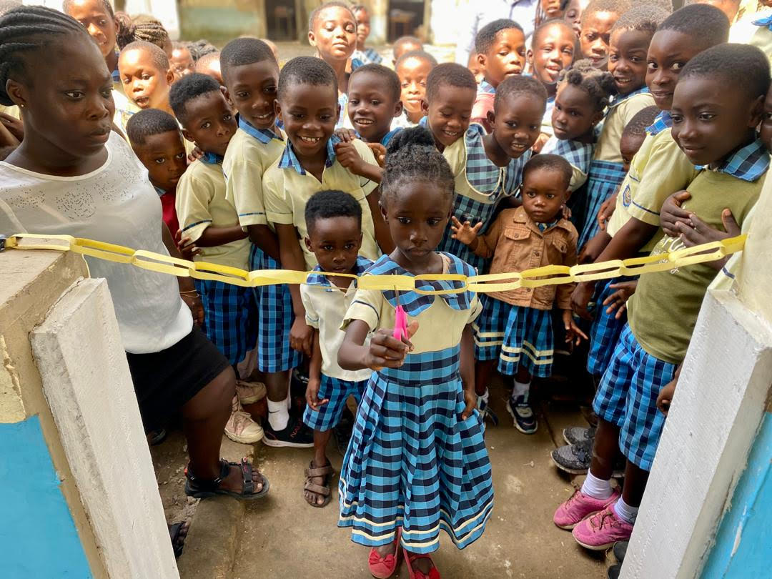A group of young African children gathered outdoors, standing behind a yellow chain barrier, with one girl cutting a pink ribbon in the center.