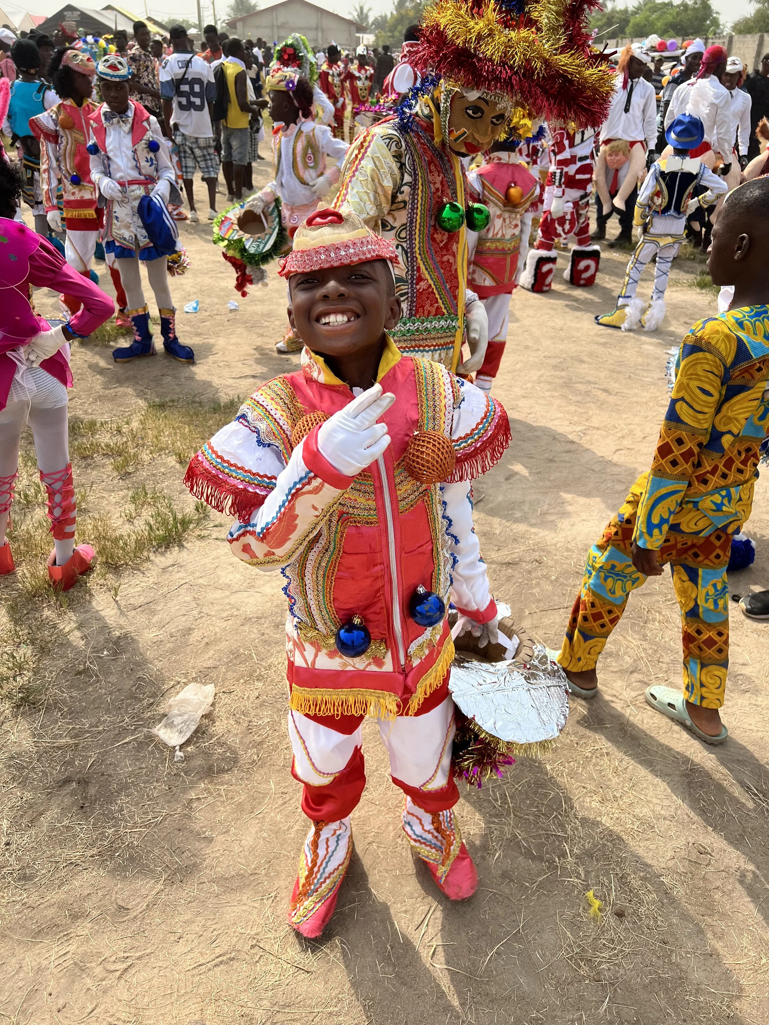 A boy dressed in colorful traditional attire, smiling and posing at an outdoor cultural event with others in similar costumes and masks in the background.