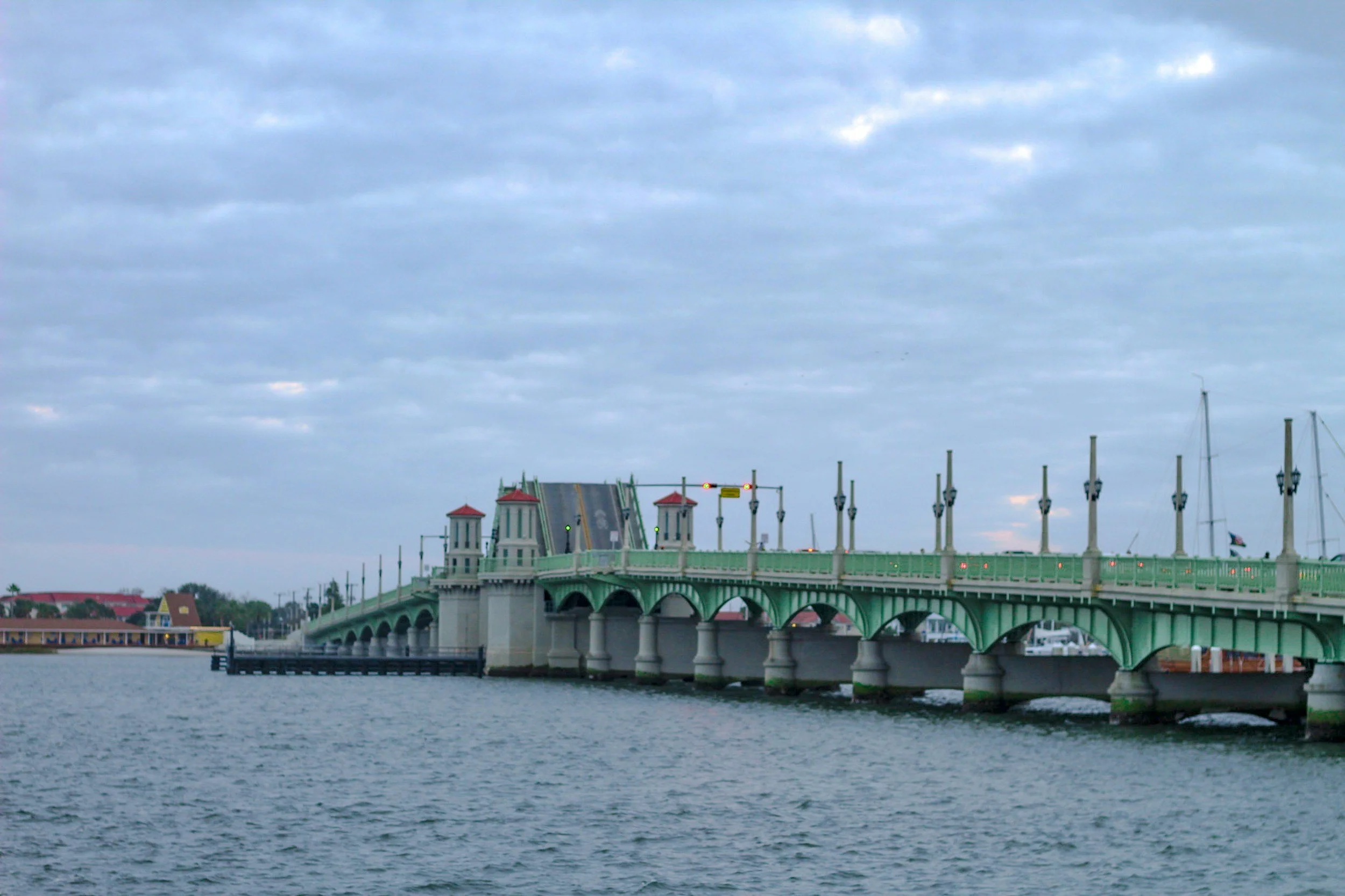 A green bridge with decorative towers over a body of water, with boat masts and a cloudy sky.